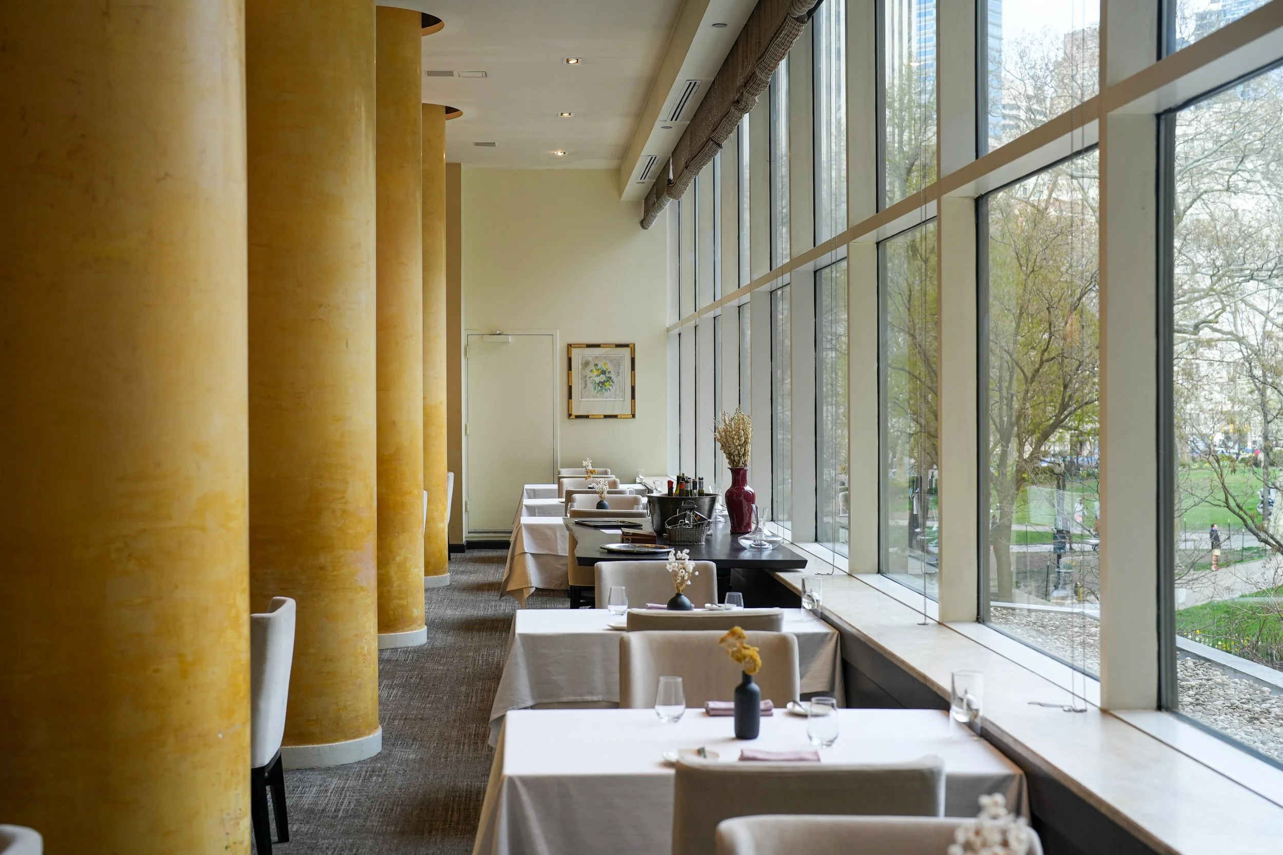 Somberly lit dining area with white-draped tables, shelves with wine bottles, and a large window overlooking a park with trees.