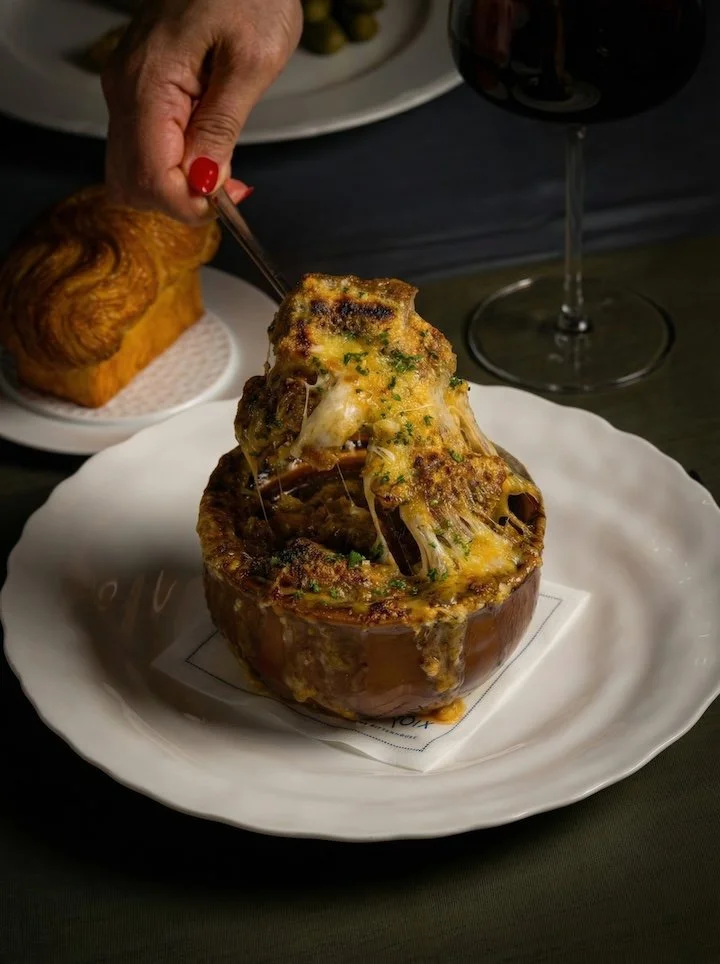 A large baked stuffed pumpkin filled with cheese, herbs, and possibly meat, being served with a fork. A glass of red wine and bread are also visible on the table.