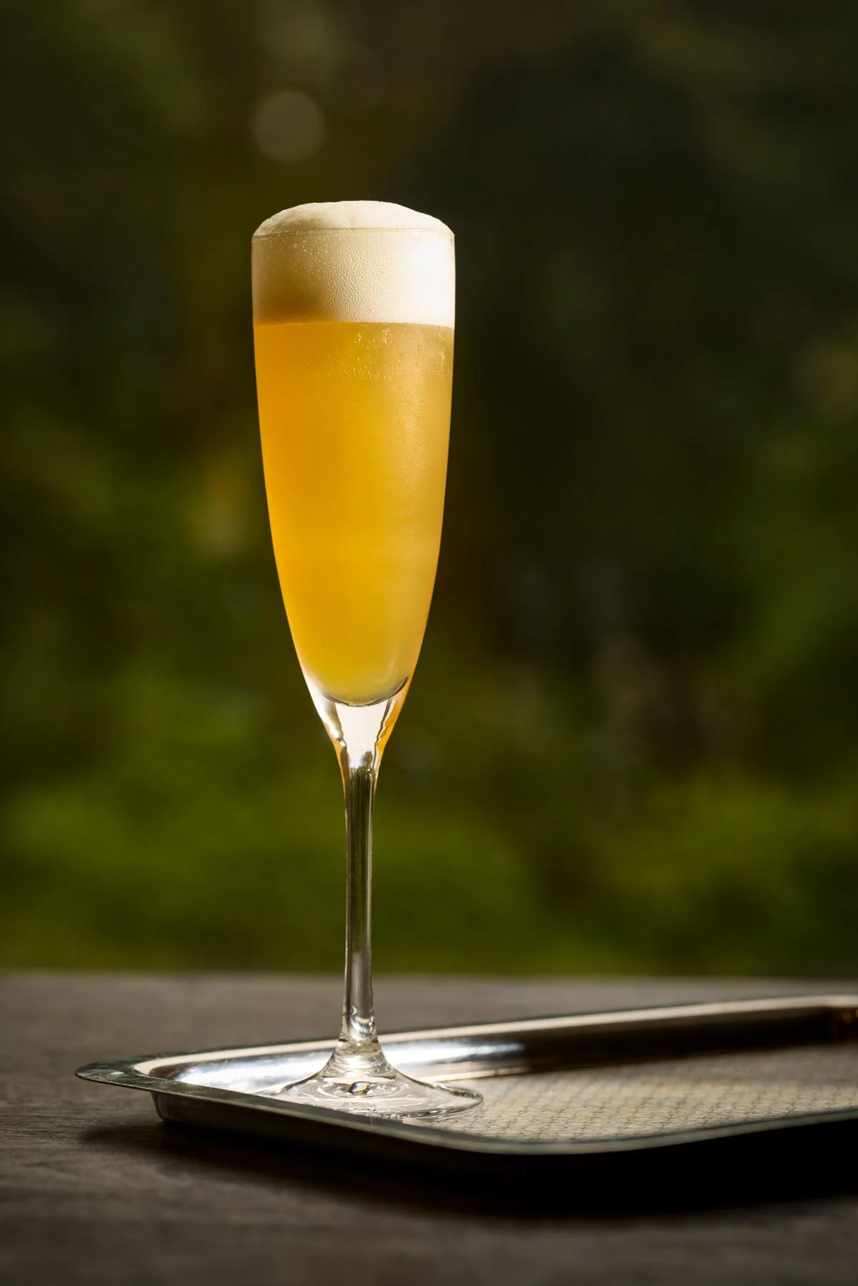 A tall flute glass filled with a yellowish beverage, topped with foam, sitting on a metallic tray with a blurred green background.