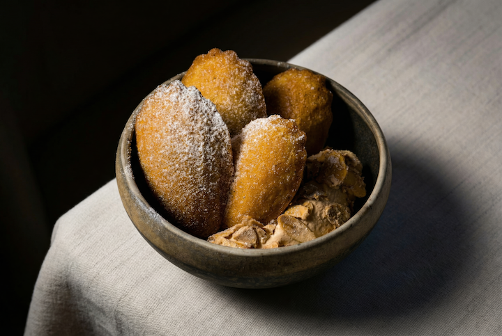 A bowl of deep-fried dough balls dusted with powdered sugar, served with a side of peanut butter or a similar spread on a white cloth