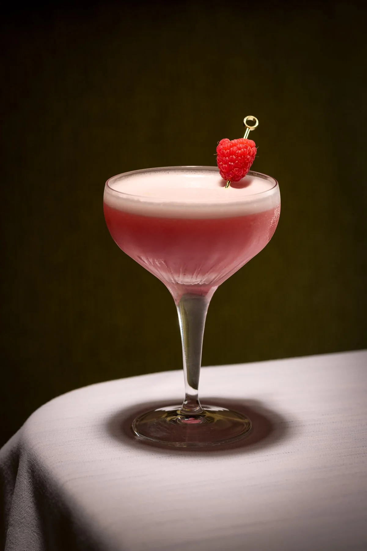 A pink cocktail in a coupe glass garnished with a raspberry on a cocktail pick.
