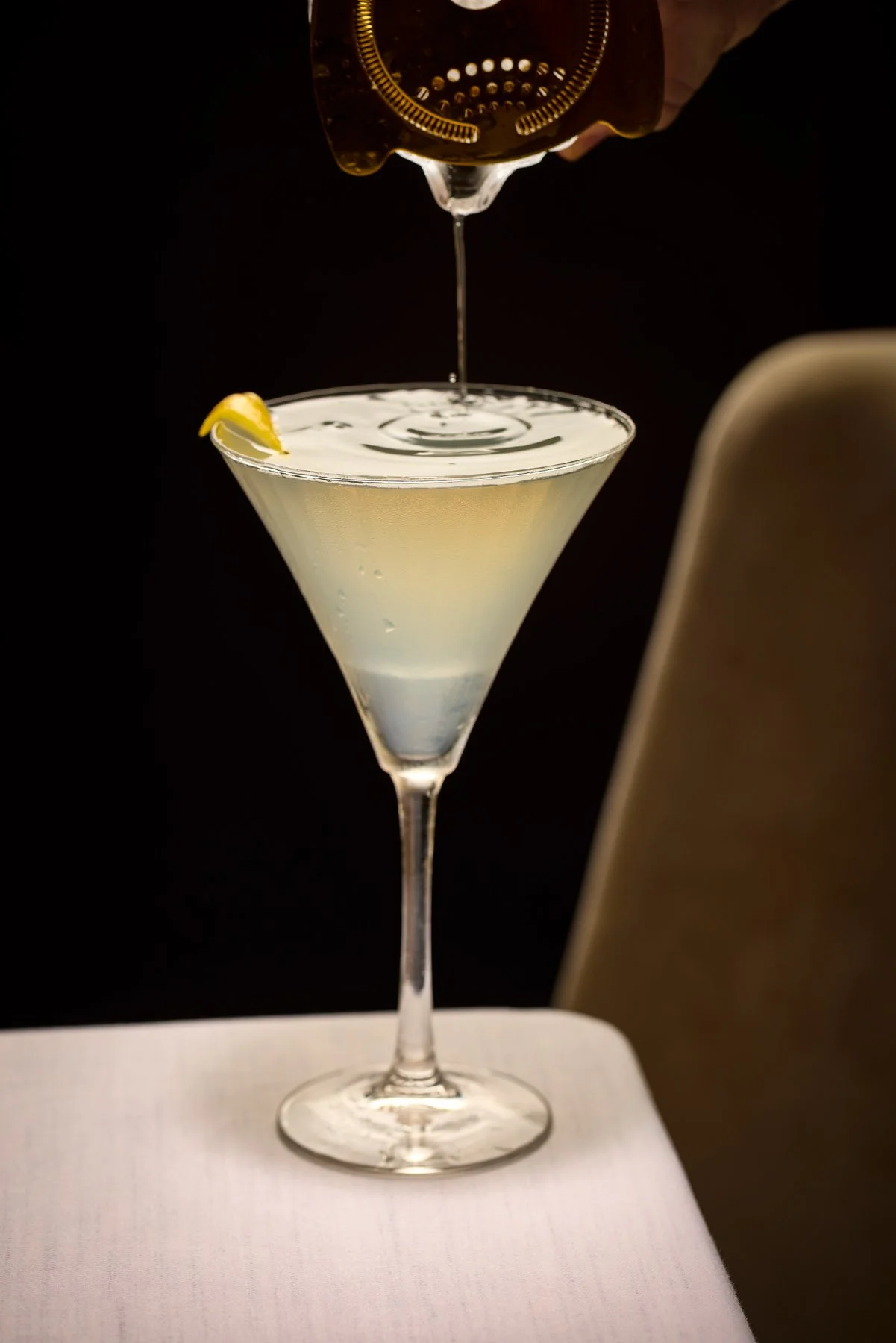 A cocktail in a martini glass with a lemon wedge garnish, being poured from a bottle over a dark background.