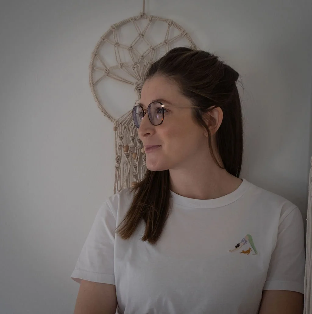 Eine junge Frau mit braunem Haar, Brille und weißem T-Shirt steht vor einer weißen Wand mit einem Wandbehang aus Makramee