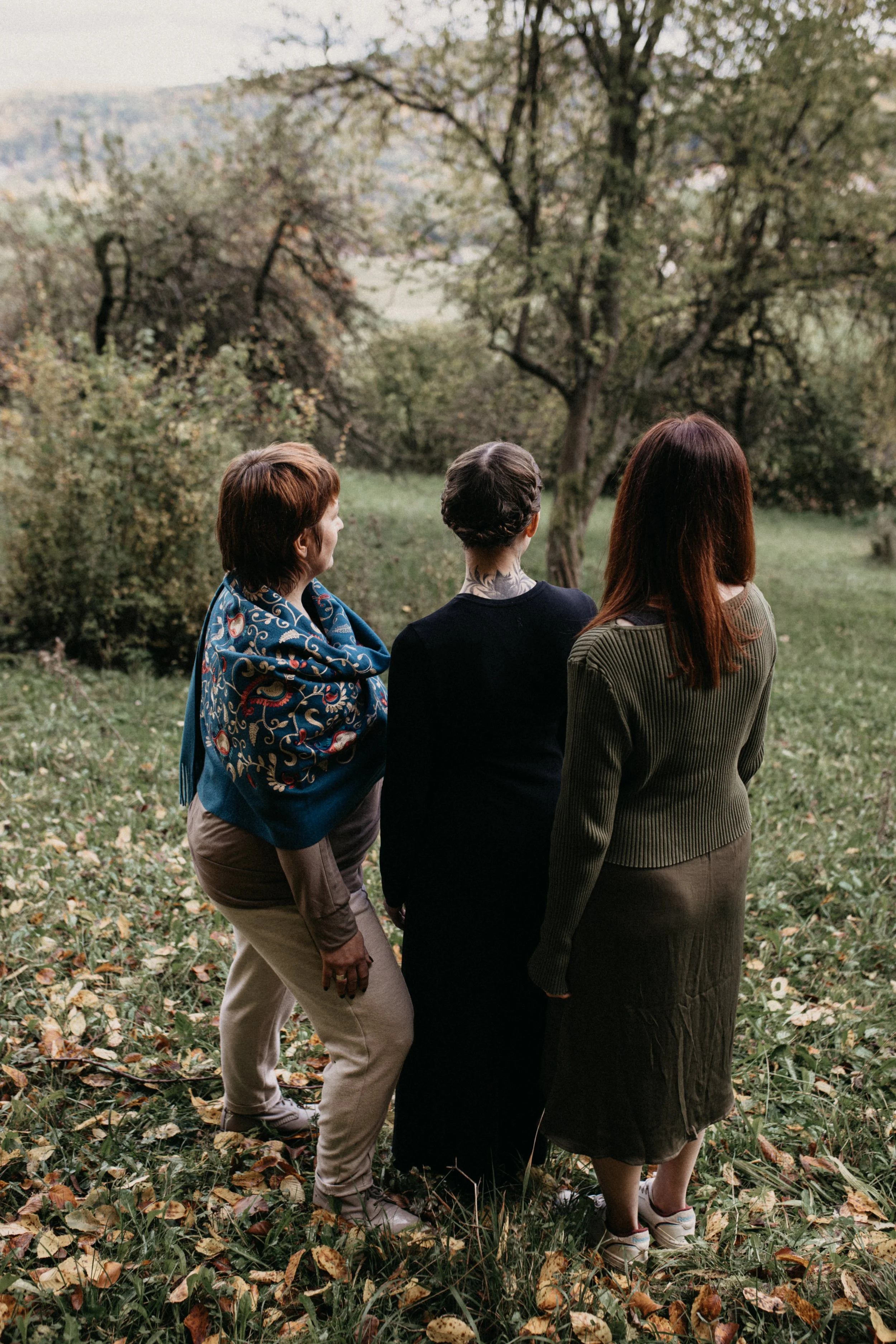 Drei Frauen stehen im Park mit herabfallenden Blättern und betrachten die Bäume in der Ferne, während der Himmel bewölkt ist.