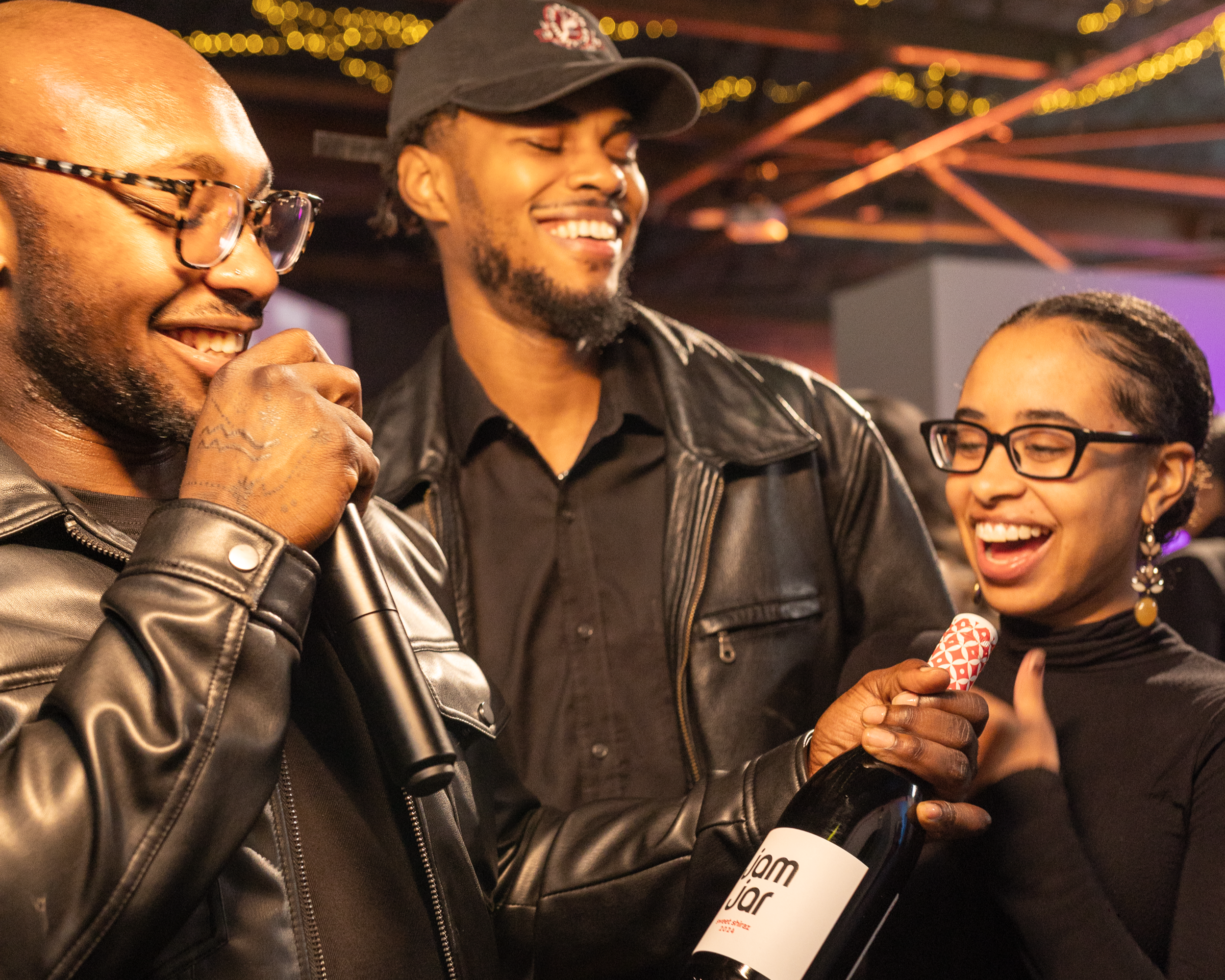Three people laughing and enjoying a moment together at a social event. One person is holding a microphone near another person, who is holding a bottle of champagne.