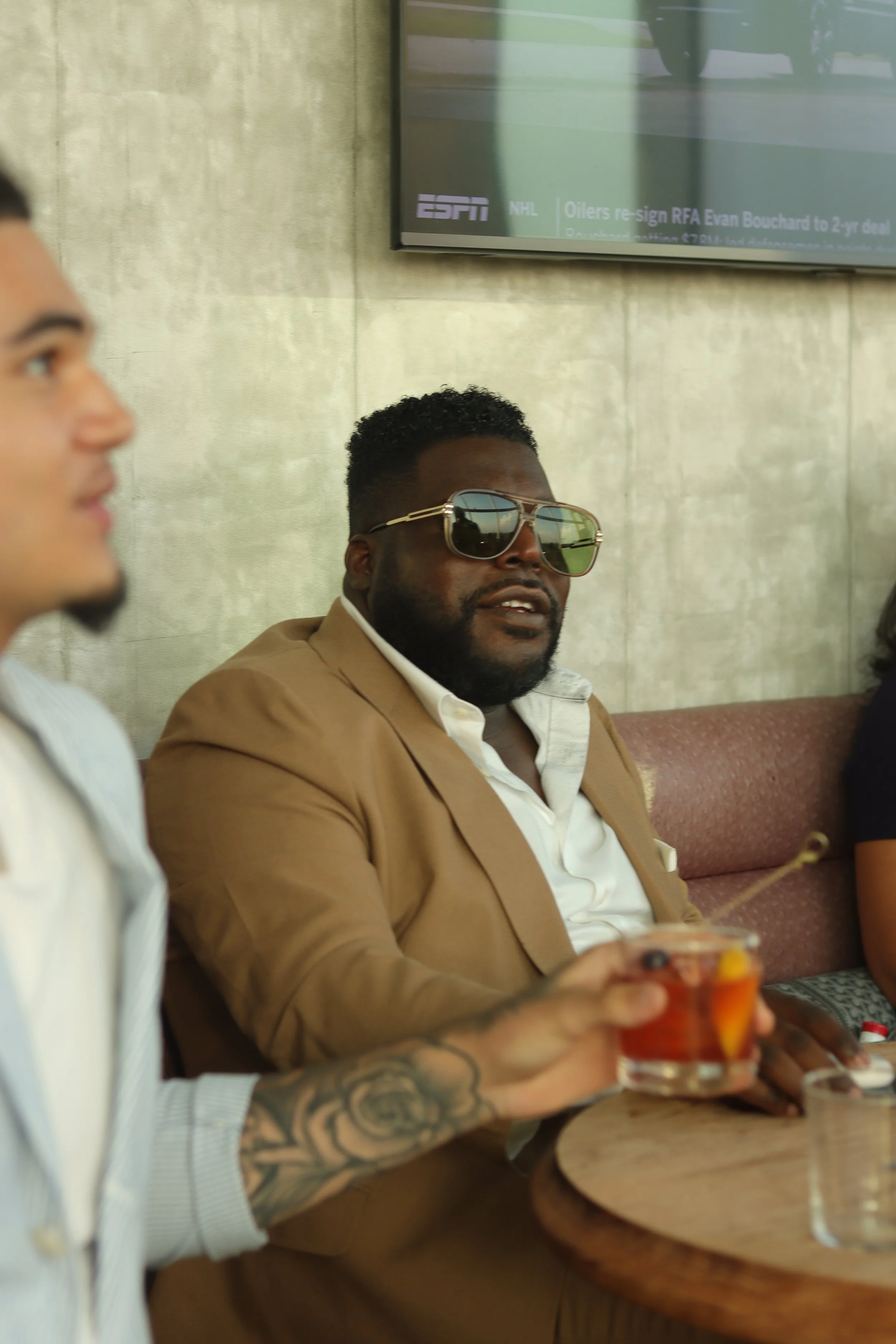 A man with sunglasses in a brown suit holding a drink at a social gathering.