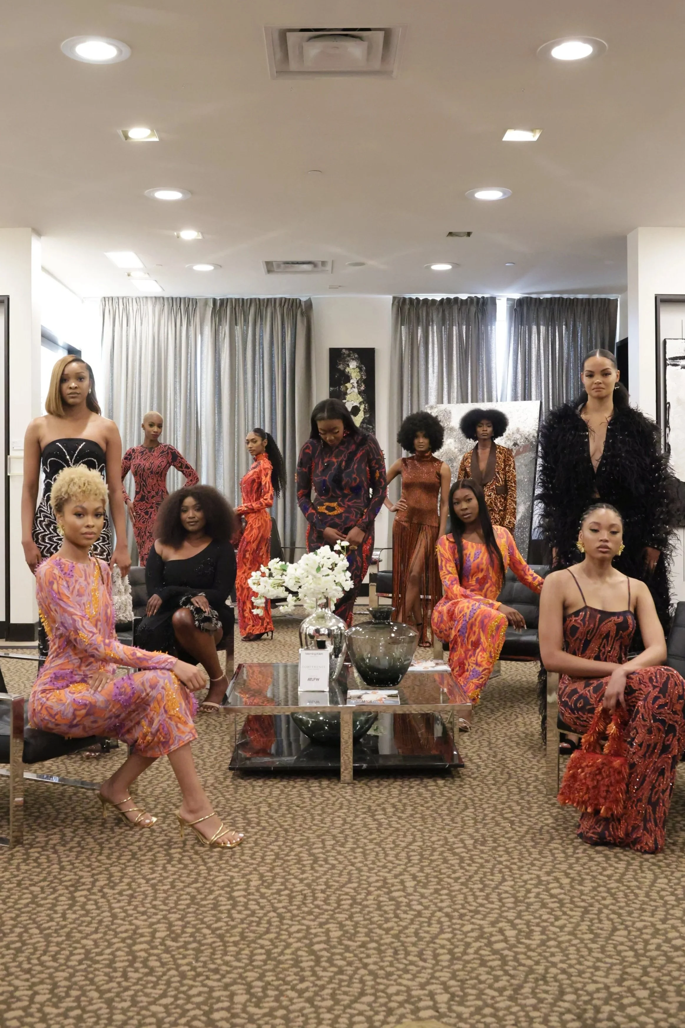 A group of women dressed in colorful and stylish outfits, standing and sitting in a modern, well-lit room with abstract artwork, a beige carpet, curtains, and a glass coffee table with decorative items.