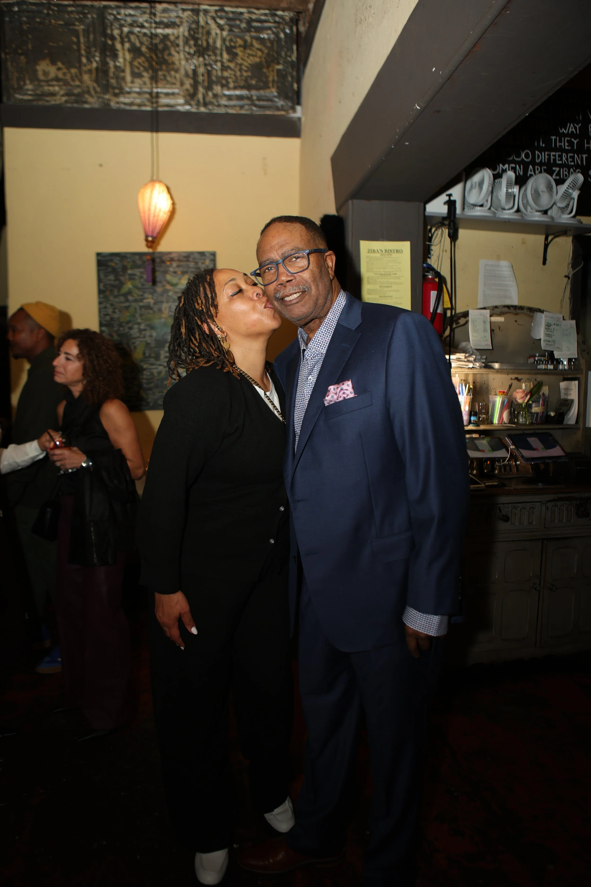 A woman kisses an older man on the cheek at a social gathering, both smiling. The man is wearing a navy blue suit with a patterned shirt and pink pocket square, while the woman is dressed in black. In the background, there are other people, a framed painting, and decorative lighting.