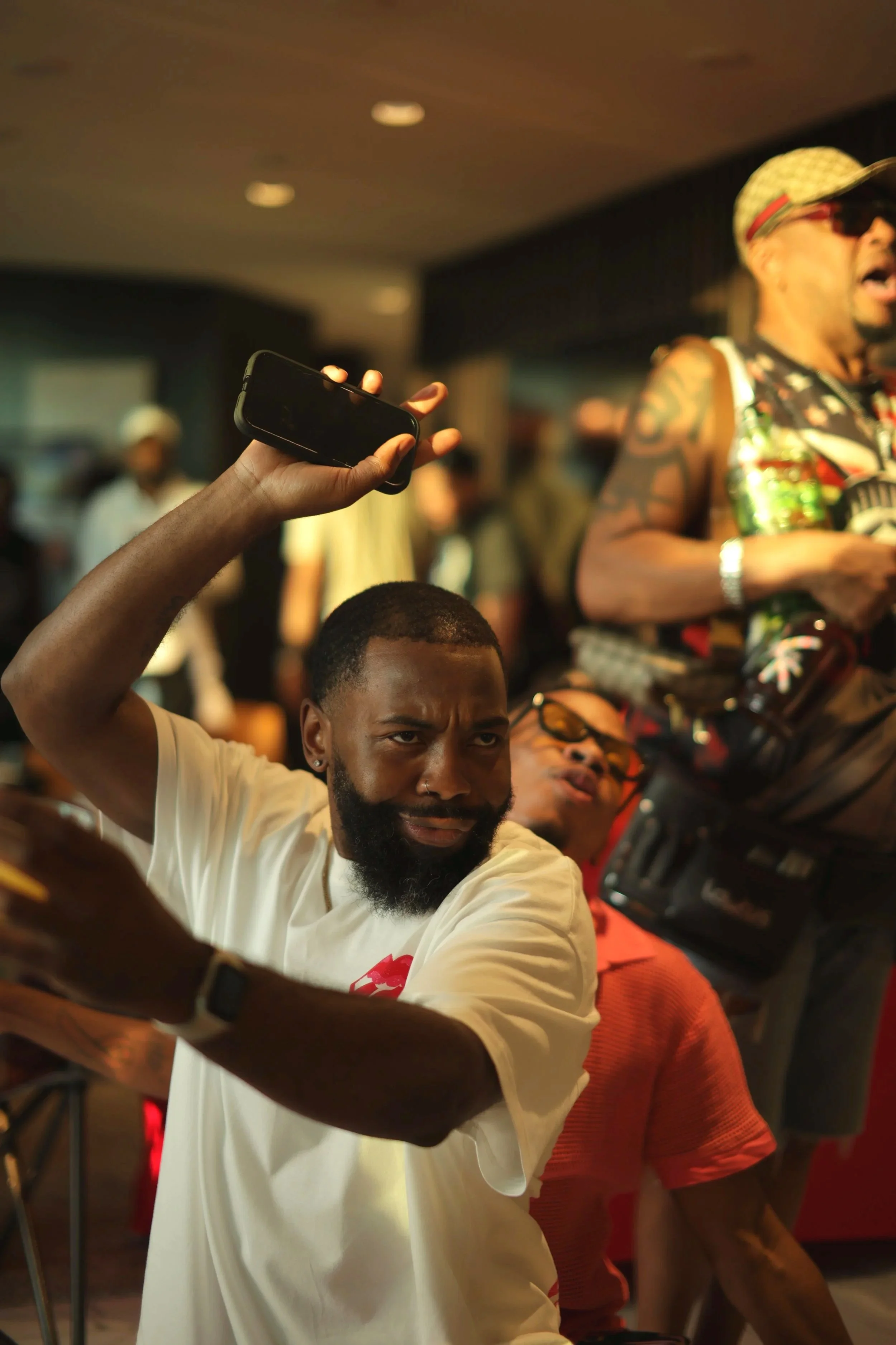 A man with a beard and mustache holds up a smartphone, appearing to take a photo or record, in a lively indoor setting with people in the background, some wearing colorful clothing and accessories.
