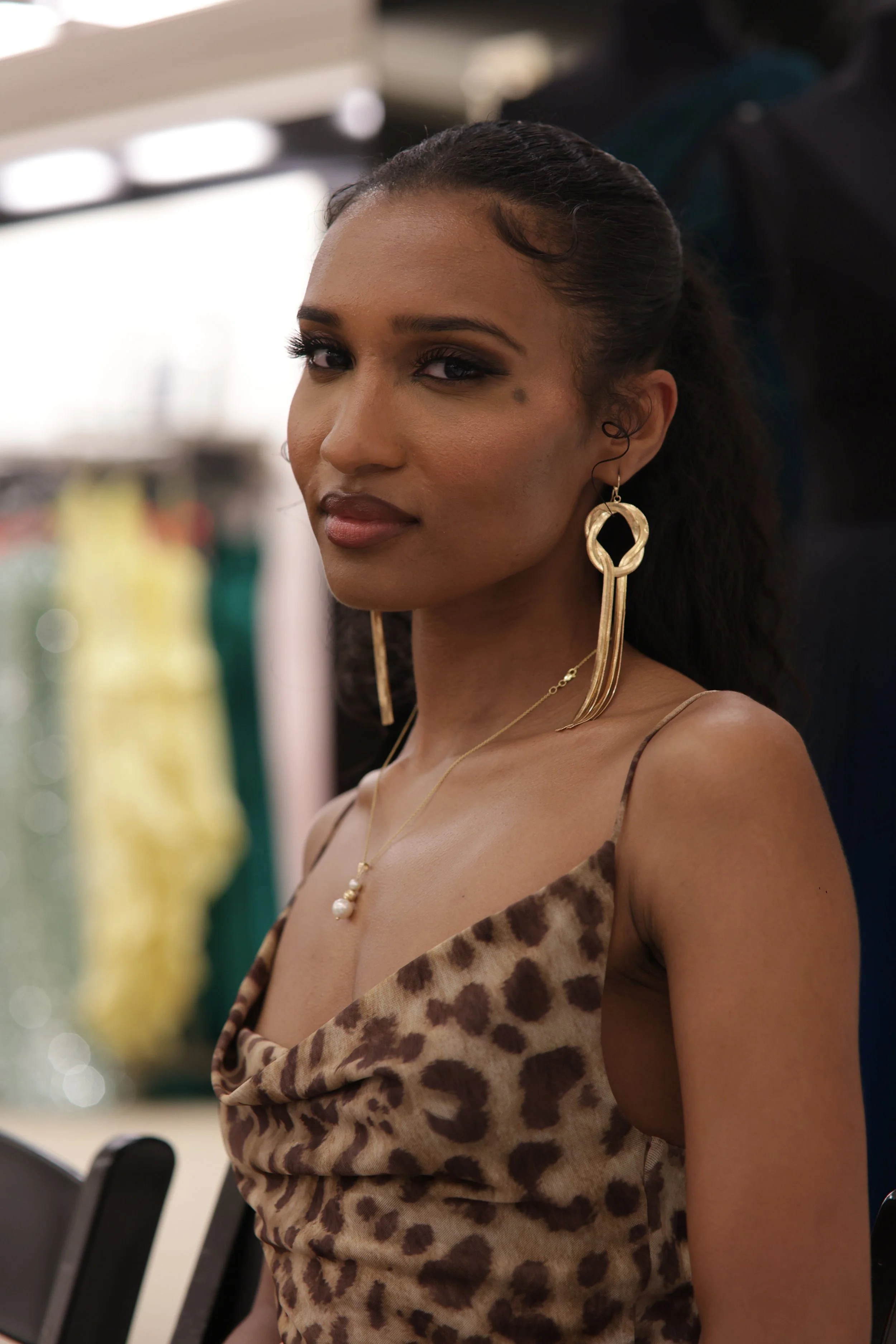 A woman with long, styled hair, wearing bold makeup, gold earrings, a gold necklace with pearls, and a leopard print dress, looking slightly to the side.