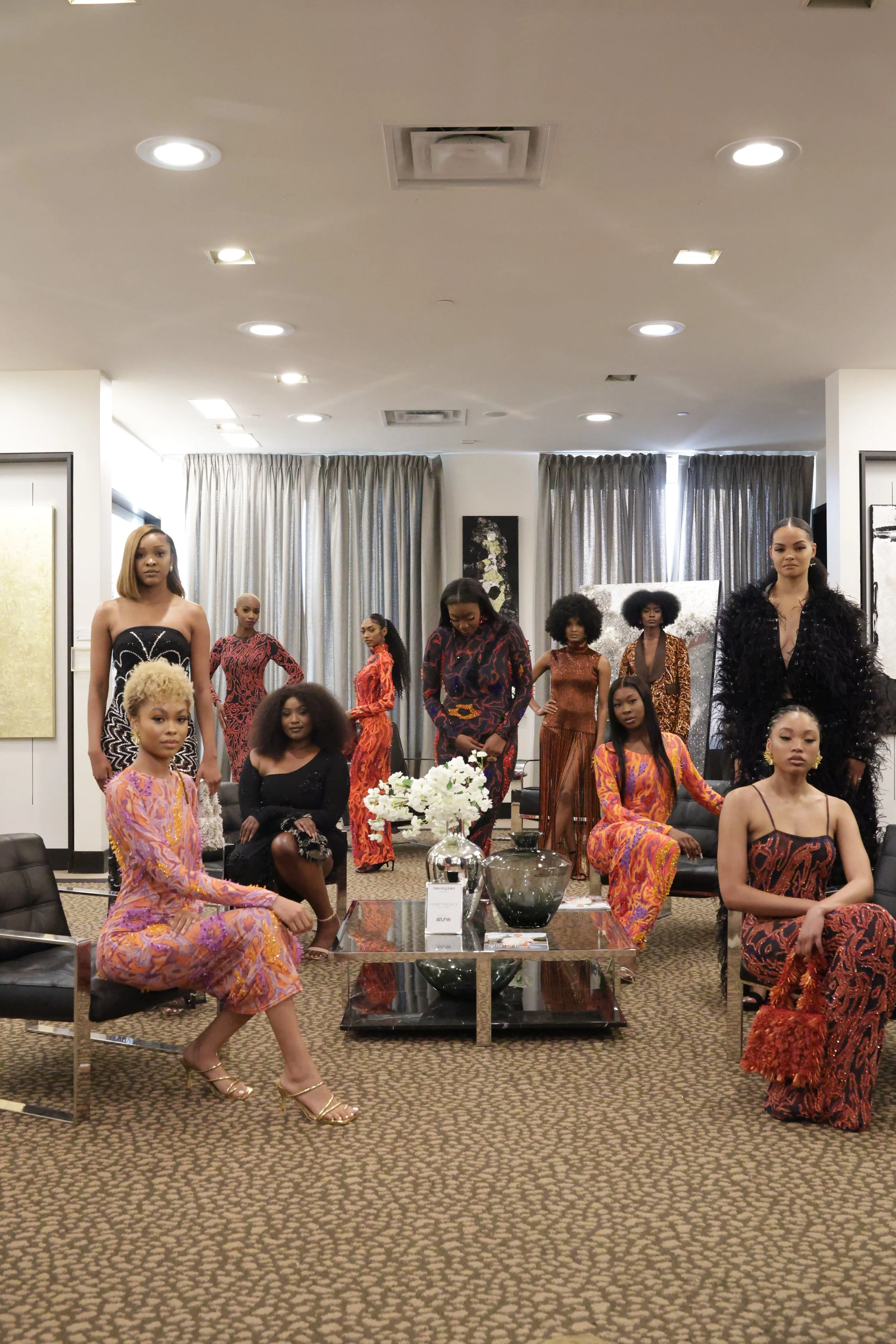 A group of women dressed in colorful and stylish outfits, standing and sitting in a modern, well-lit room with abstract artwork, a beige carpet, curtains, and a glass coffee table with decorative items.