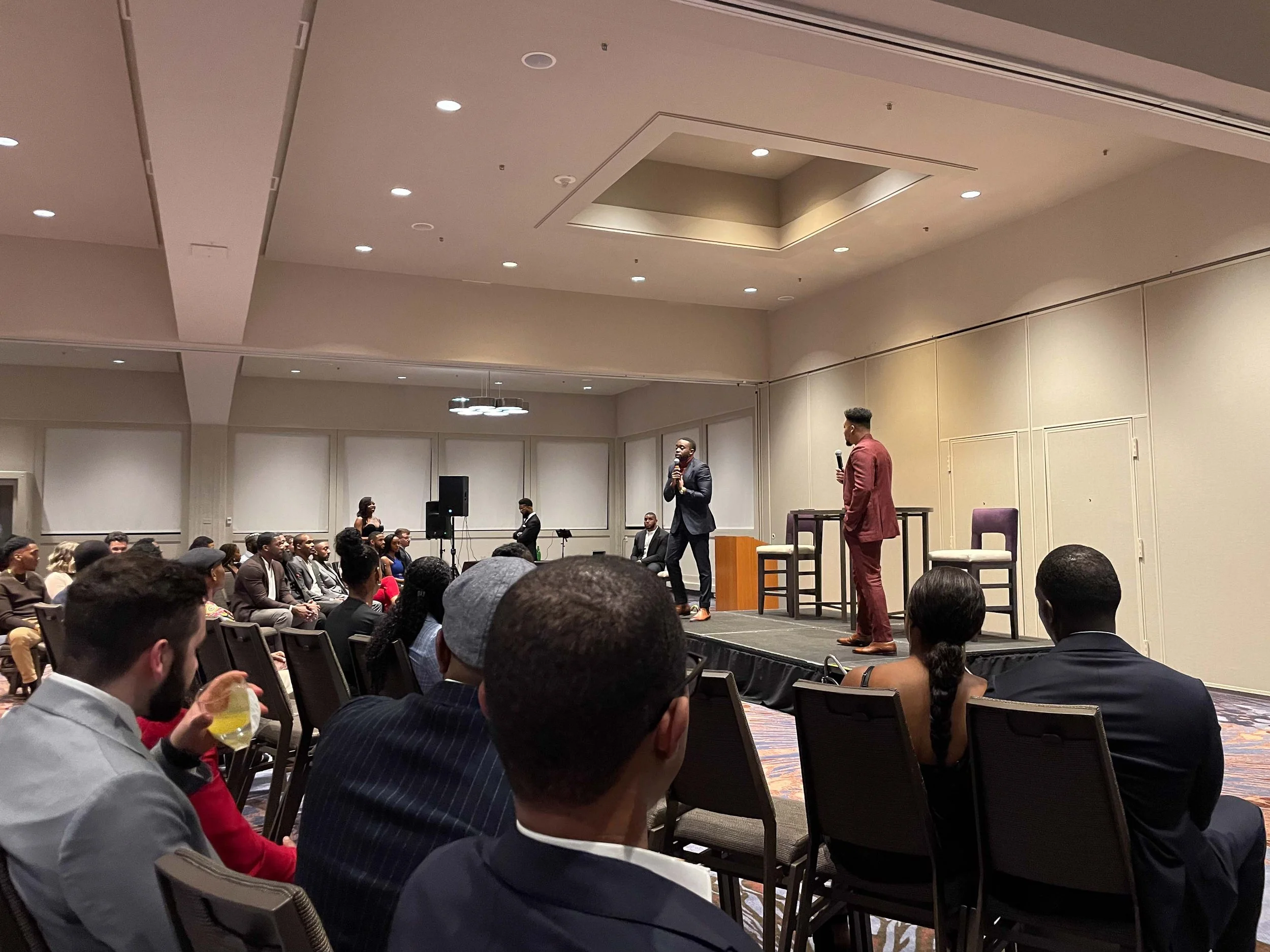 A conference or seminar with two men speaking on stage, an audience seated facing them, in a large, well-lit room with beige walls and ceiling lights.