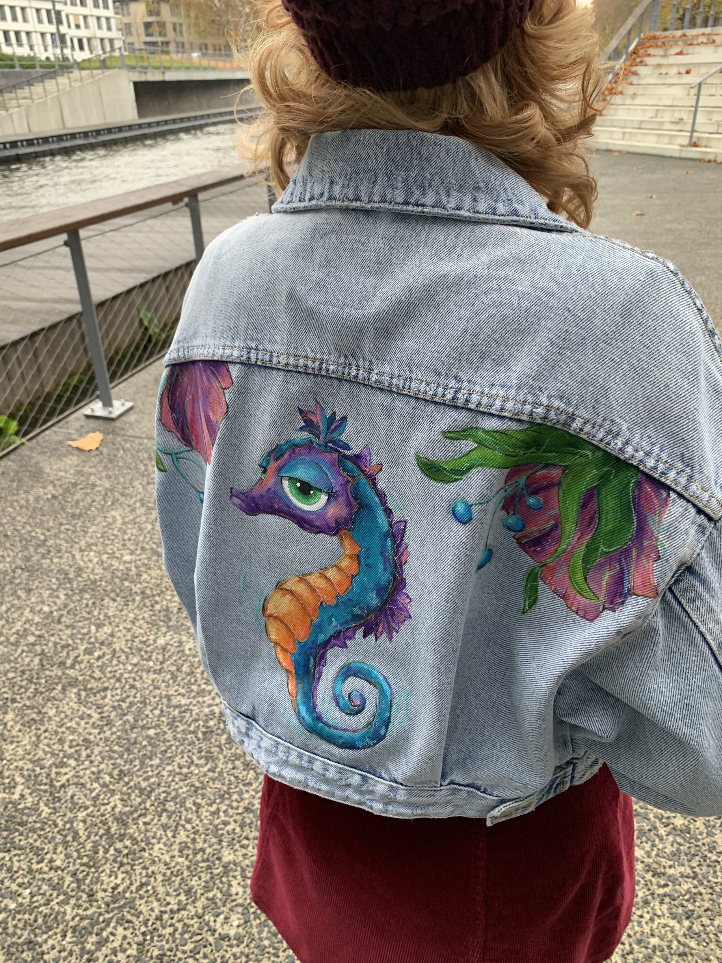 Back of a person wearing a denim jacket with a colorful painted seahorse, flowers, and leaves design, standing near water and stairs outdoors.
