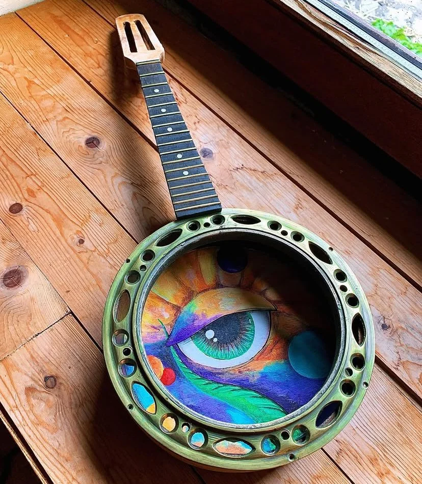 Circular object resembling a musical instrument with an artistic eye painting inside, connected to a guitar neck, placed on wooden floor near a window.