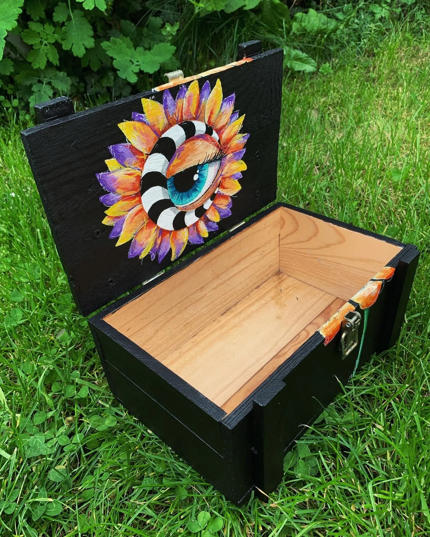 Open black wooden box with a hand-painted eye and sunflower petals design inside the lid, placed on grass.