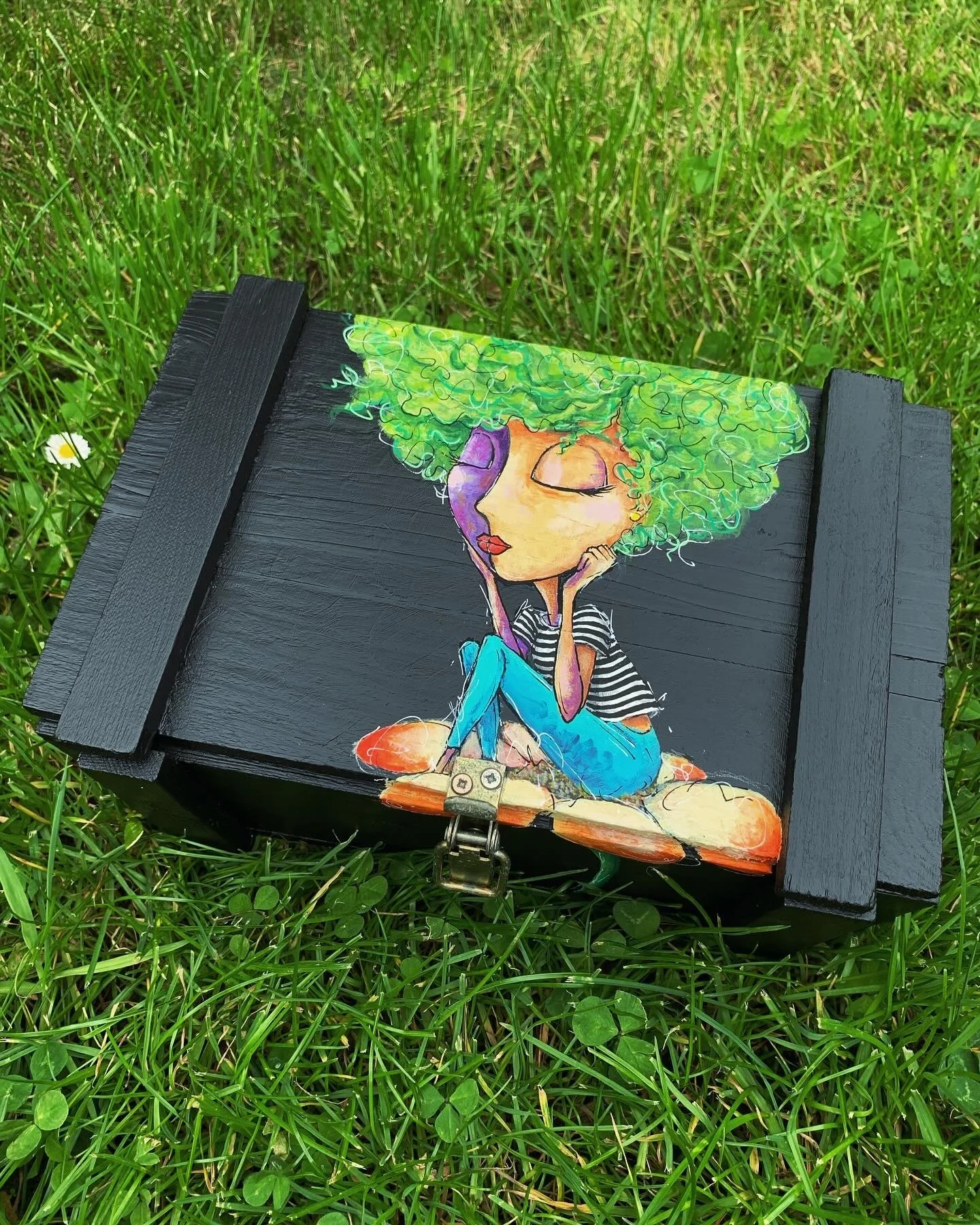 Hand-painted wooden box with a colorful illustration of a girl with curly green hair, sitting cross-legged with eyes closed, wearing a striped shirt and blue pants, on a grassy surface.