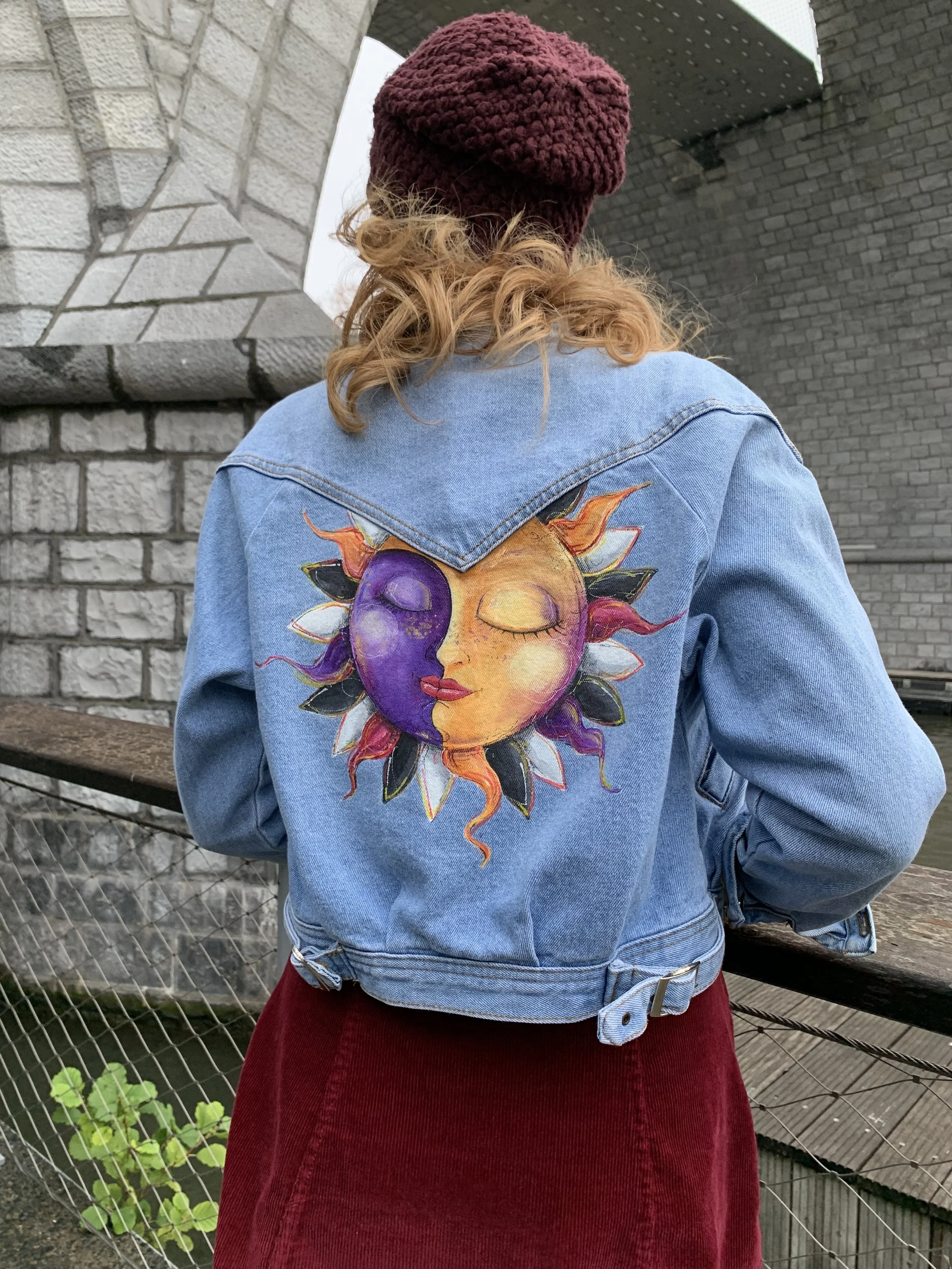 Back view of a person wearing a denim jacket with a painted sun and moon face on the back, a maroon knit beanie, and a red skirt, standing outdoors near a stone bridge and fence.