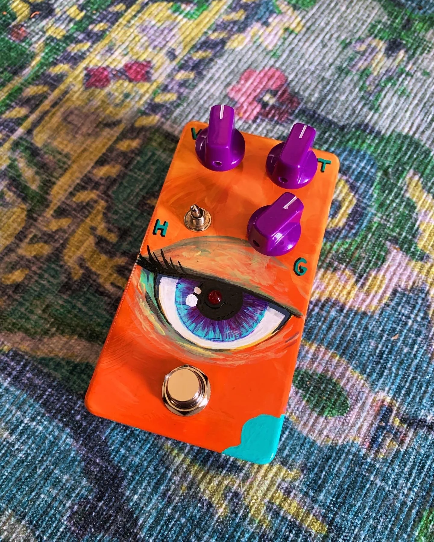 A colorful guitar effects pedal with an eye illustration and three purple knobs on a multicolored textile surface.