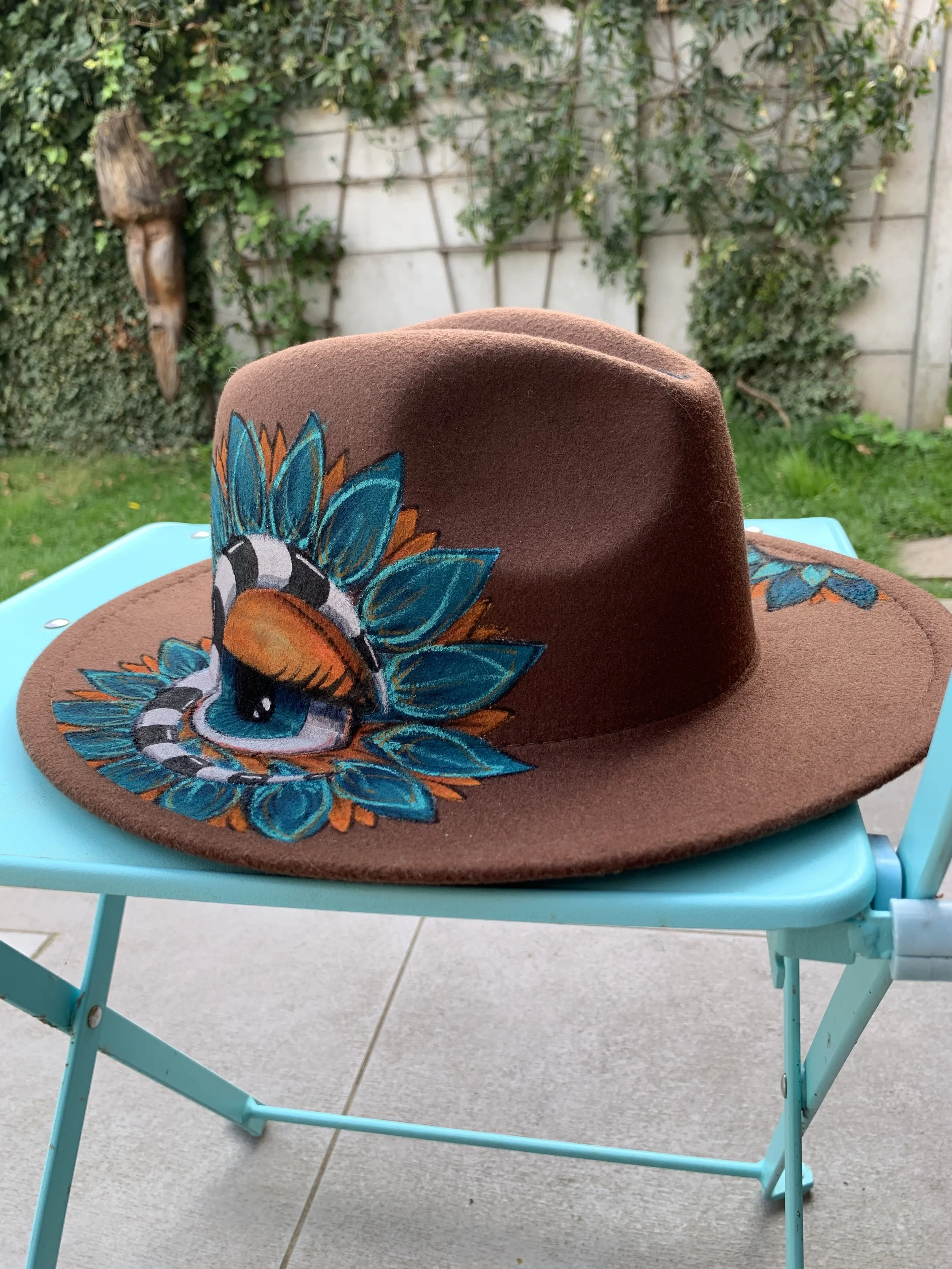 A brown felt hat with colorful embroidered feathers and a snake design, placed on a turquoise outdoor chair with a garden background.