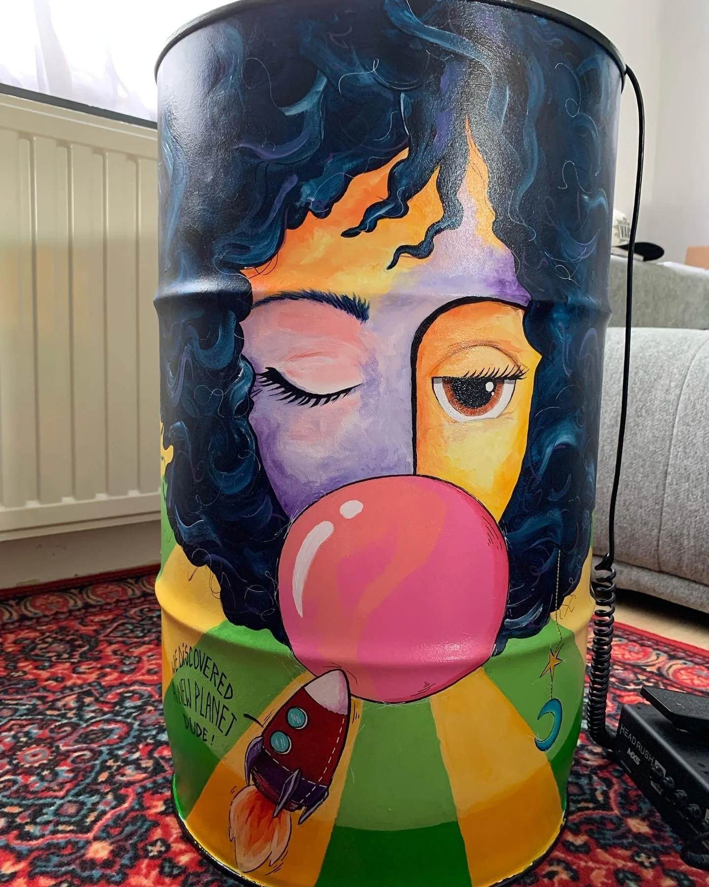 Colorful painted drum featuring a woman's face with curly blue hair, one eye closed, one eye open, blowing a pink bubble gum, with rainbow background and a small rocket ship.