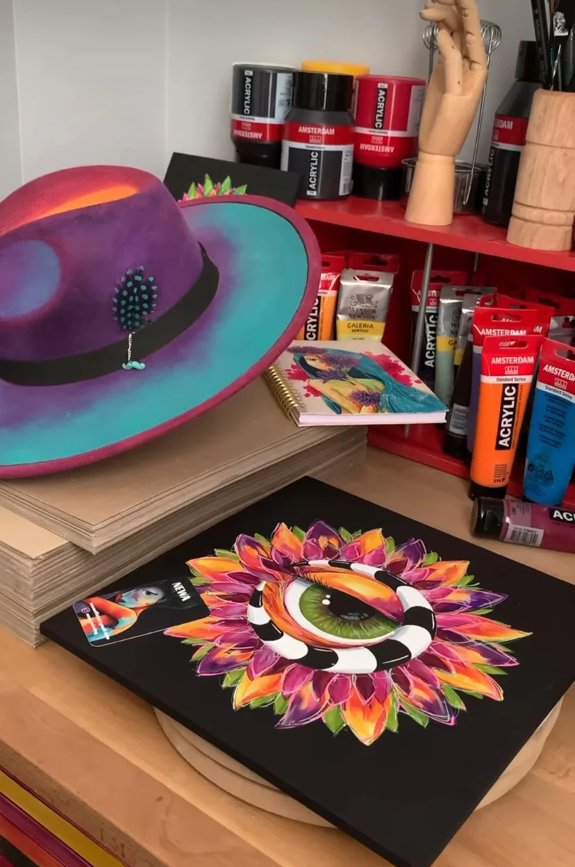 A workspace with a colorful painted hat, a matching artwork with a large eye and flowers, acrylic paint tubes, a spiral notebook with a horse painting, and artist tools and supplies.