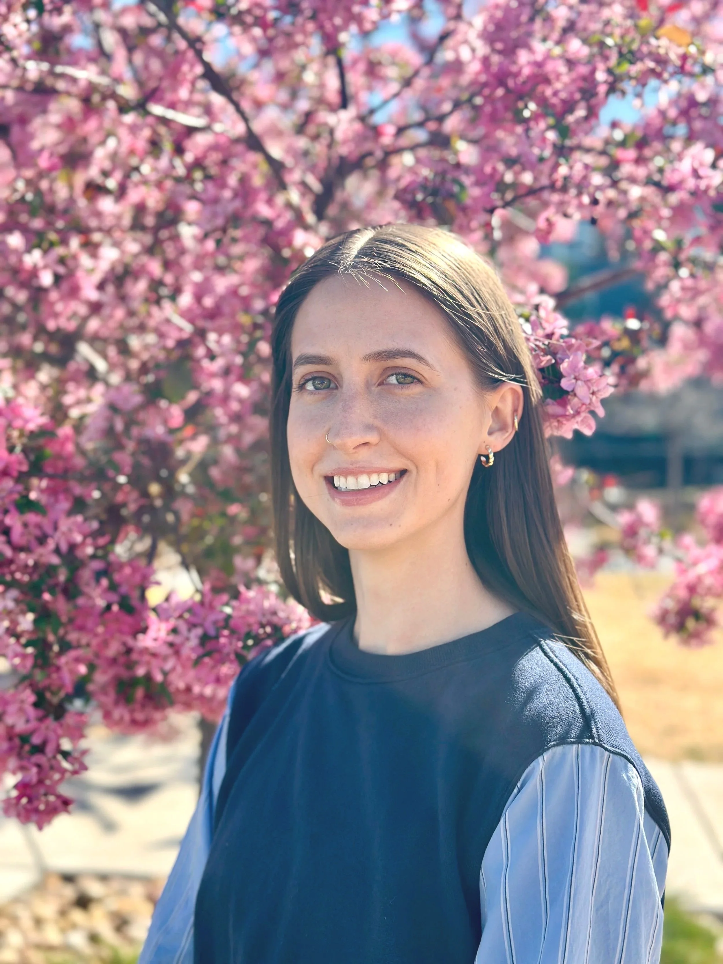 A young woman with long brown hair, light skin, and jewelry, smiling outdoors in front of a blooming pink cherry blossom tree.