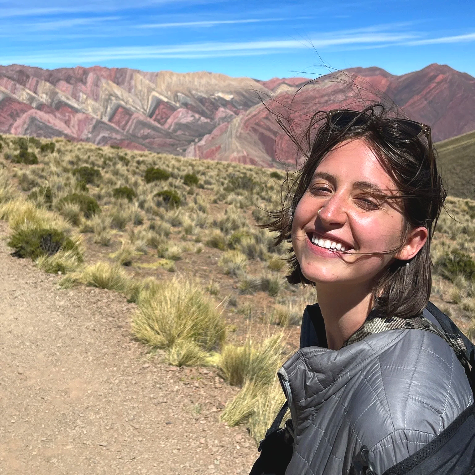 Young woman smiling outdoors in a desert landscape with colorful mountains in the background, wearing a gray jacket and carrying a backpack.
