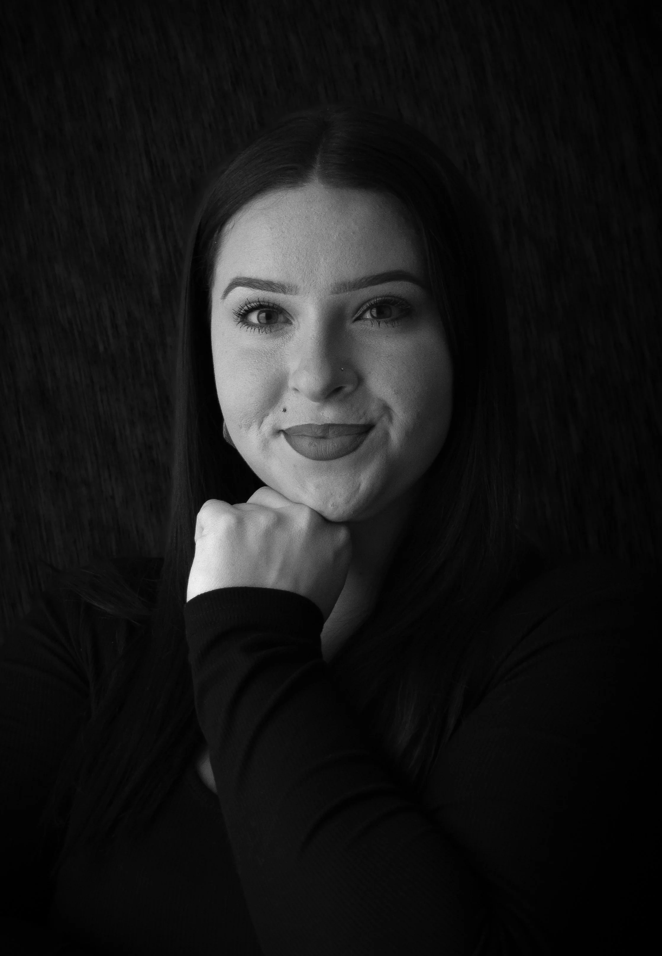Black and white portrait of a young woman with long dark hair, resting her chin on her hand, smiling softly at the camera.