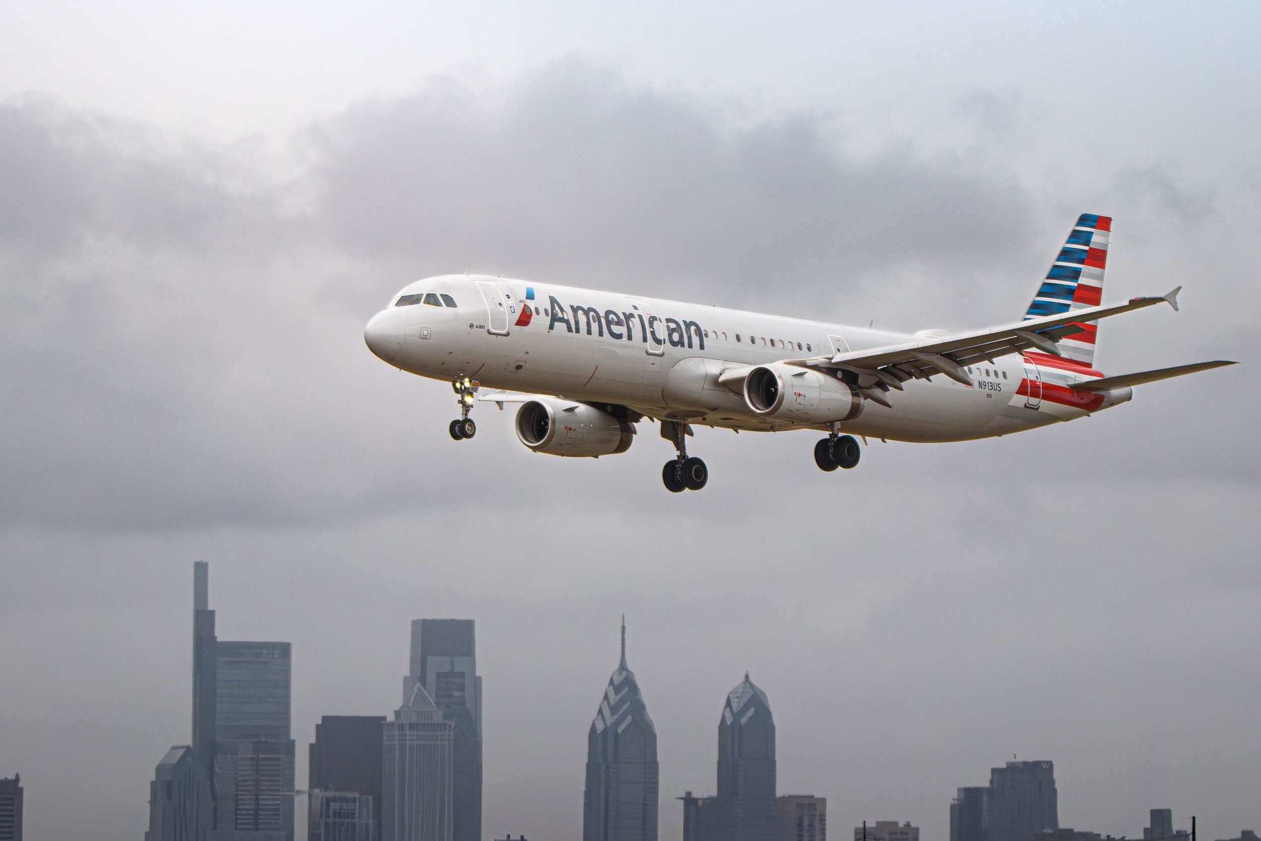 An American Airlines airplane flying over a city skyline with gray clouds in the sky.