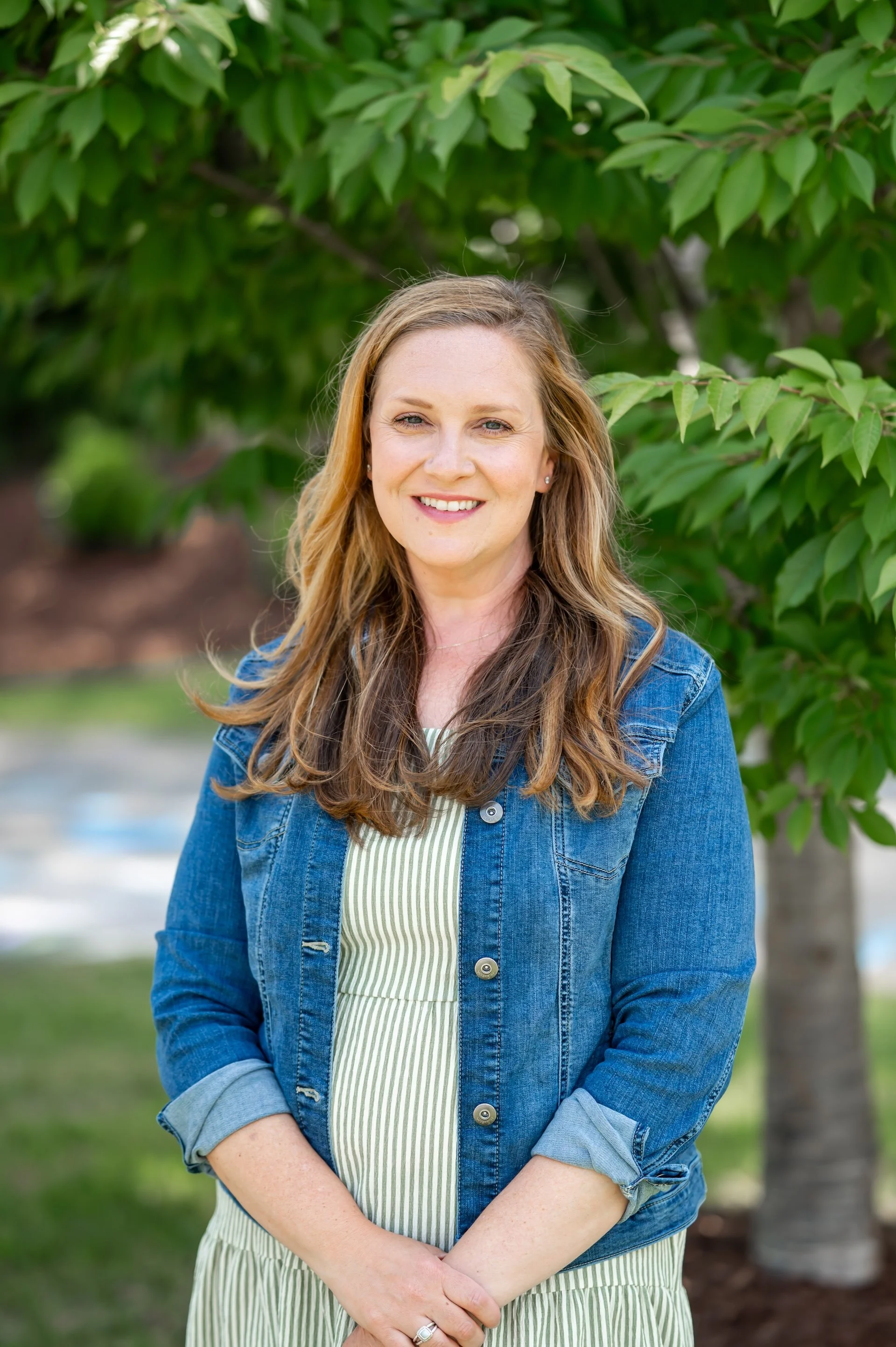 A woman with long, wavy light brown hair, wearing a denim jacket and a green and white striped dress, standing outside in front of a lush green tree, smiling at the camera.