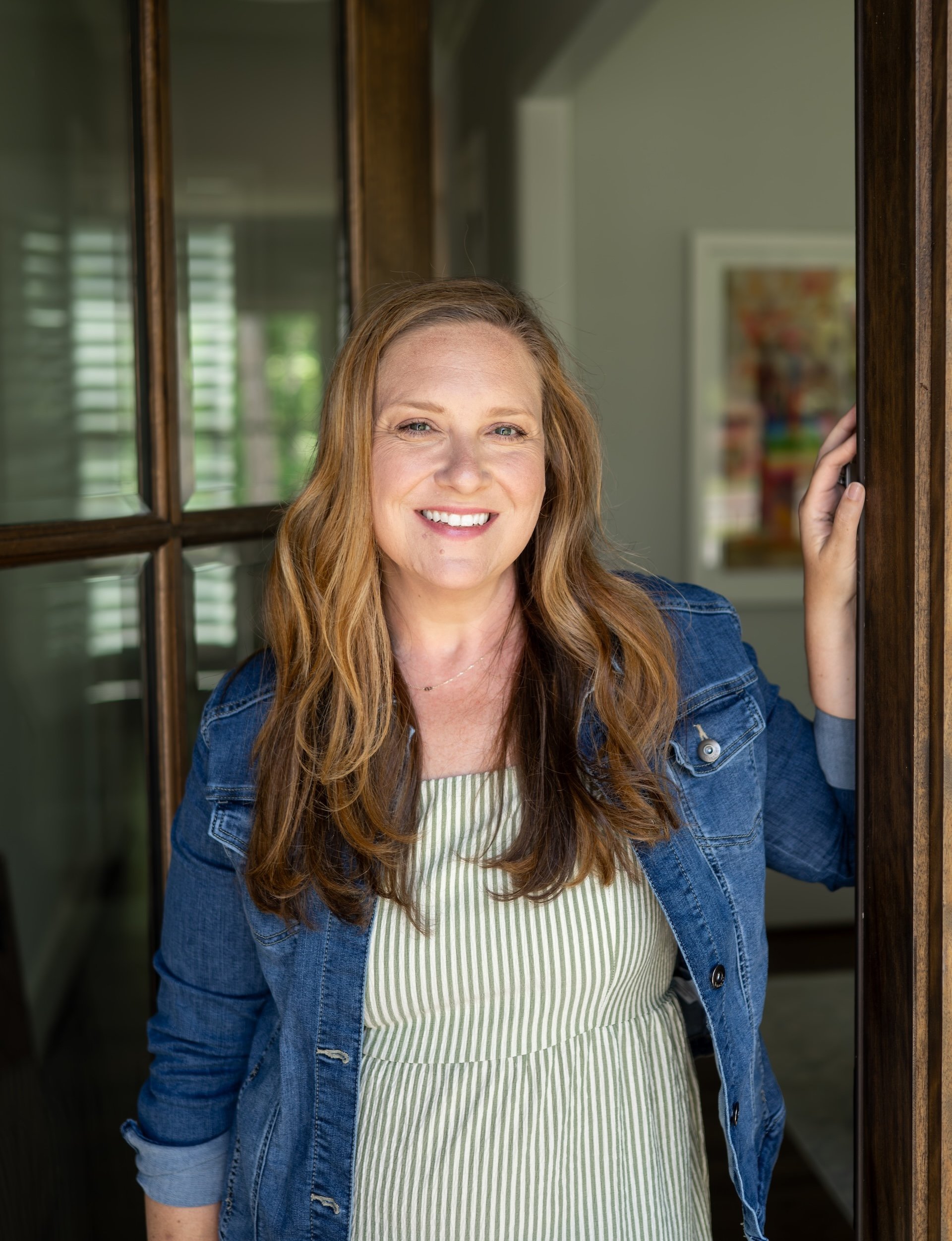 A woman with long reddish hair smiling and looking at the camera, standing next to a wooden door frame, wearing a denim jacket over a green and white striped dress.