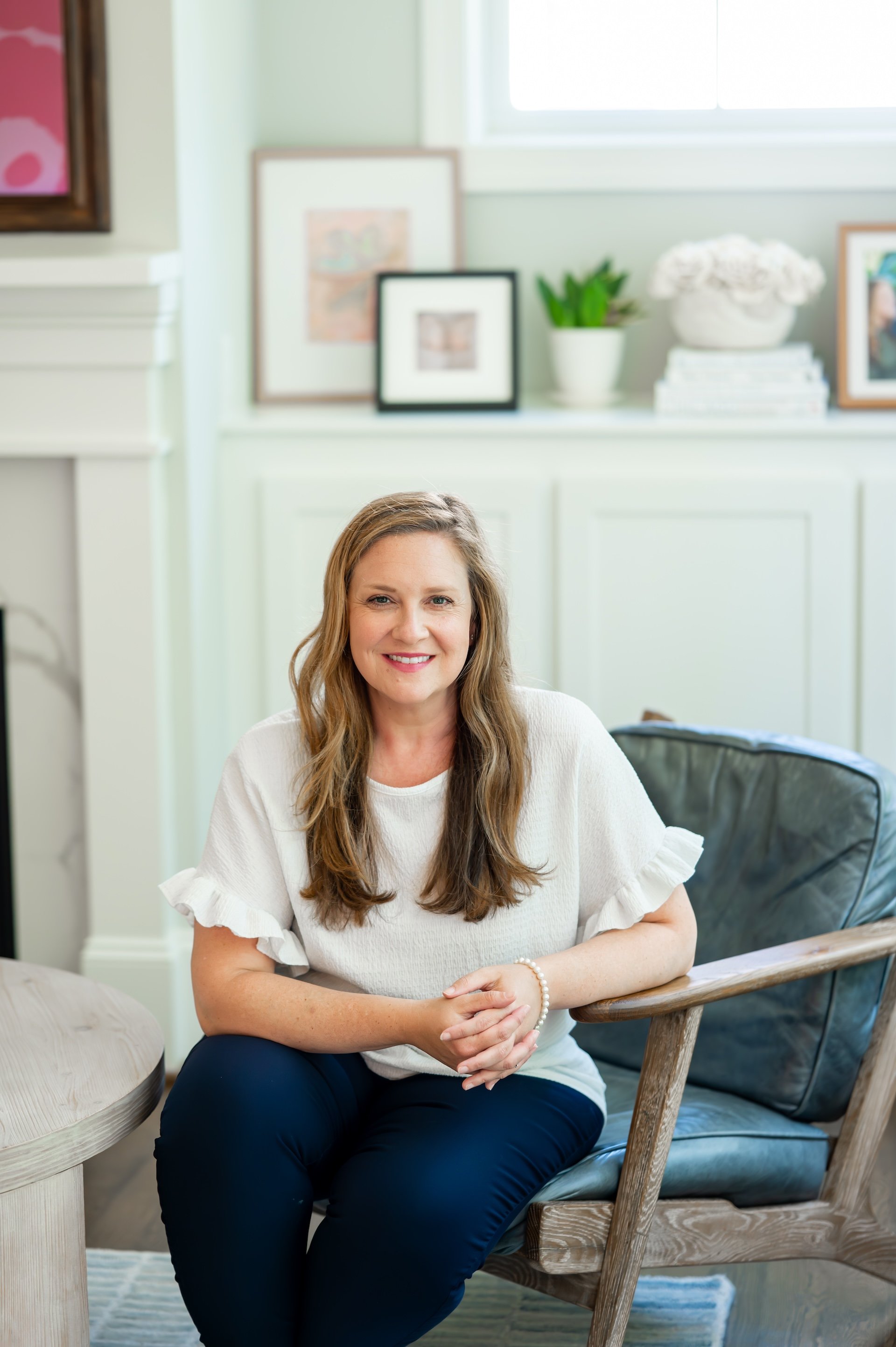 A woman with long light brown hair, smiling, wearing a white top with ruffled sleeves and black pants, sitting on a wooden chair in a brightly lit room with light-colored walls, framed artwork on the wall, a potted plant, a stack of books, and a bouquet of white flowers on a shelf in the background.