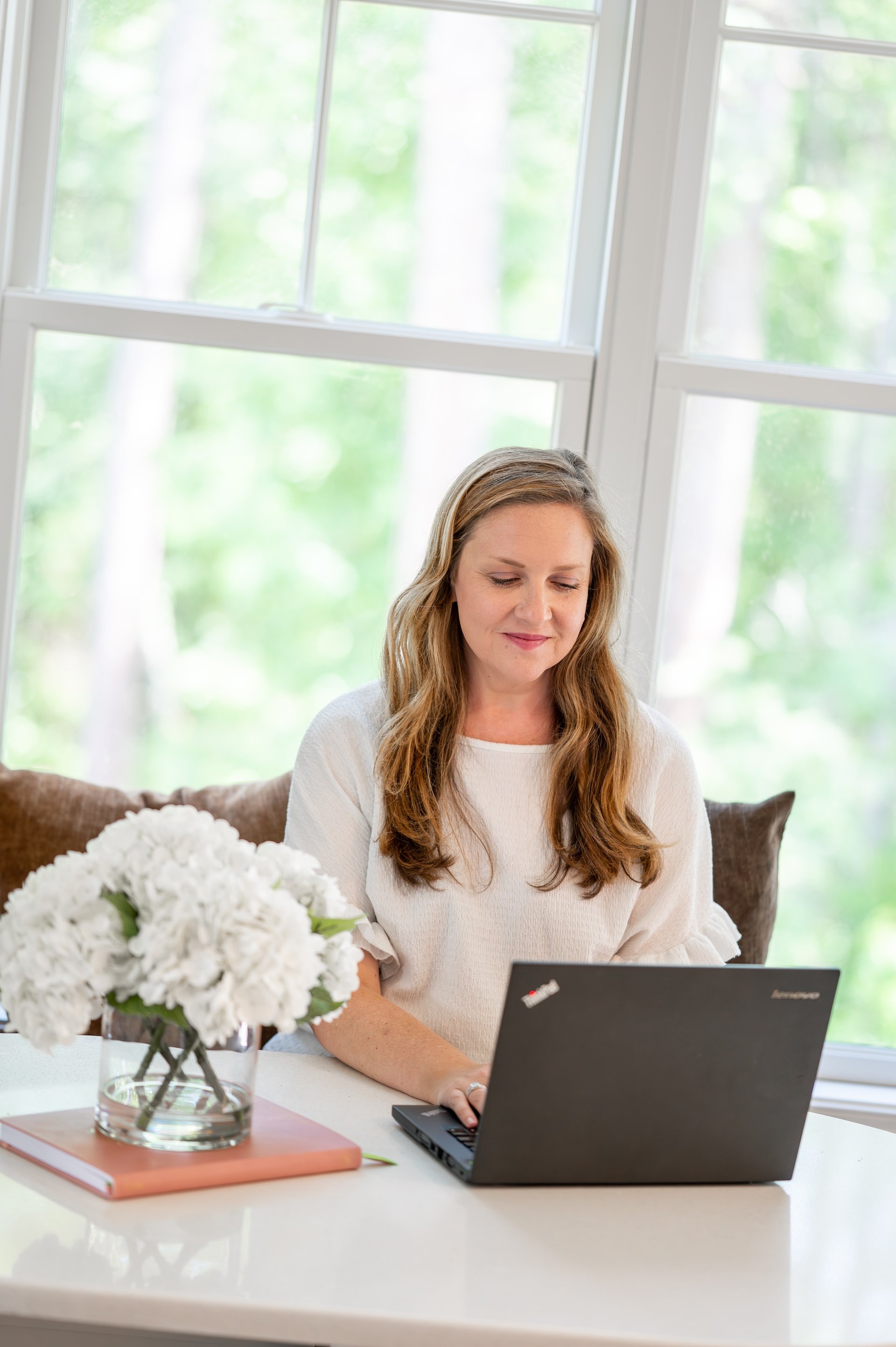 A woman with long, wavy, light brown hair sitting at a white table, working on a laptop. There is a bouquet of white flowers in a glass vase, a stack of pink books, and a closed laptop with a Lenovo logo in front of her. Large windows with greenery outside are in the background.