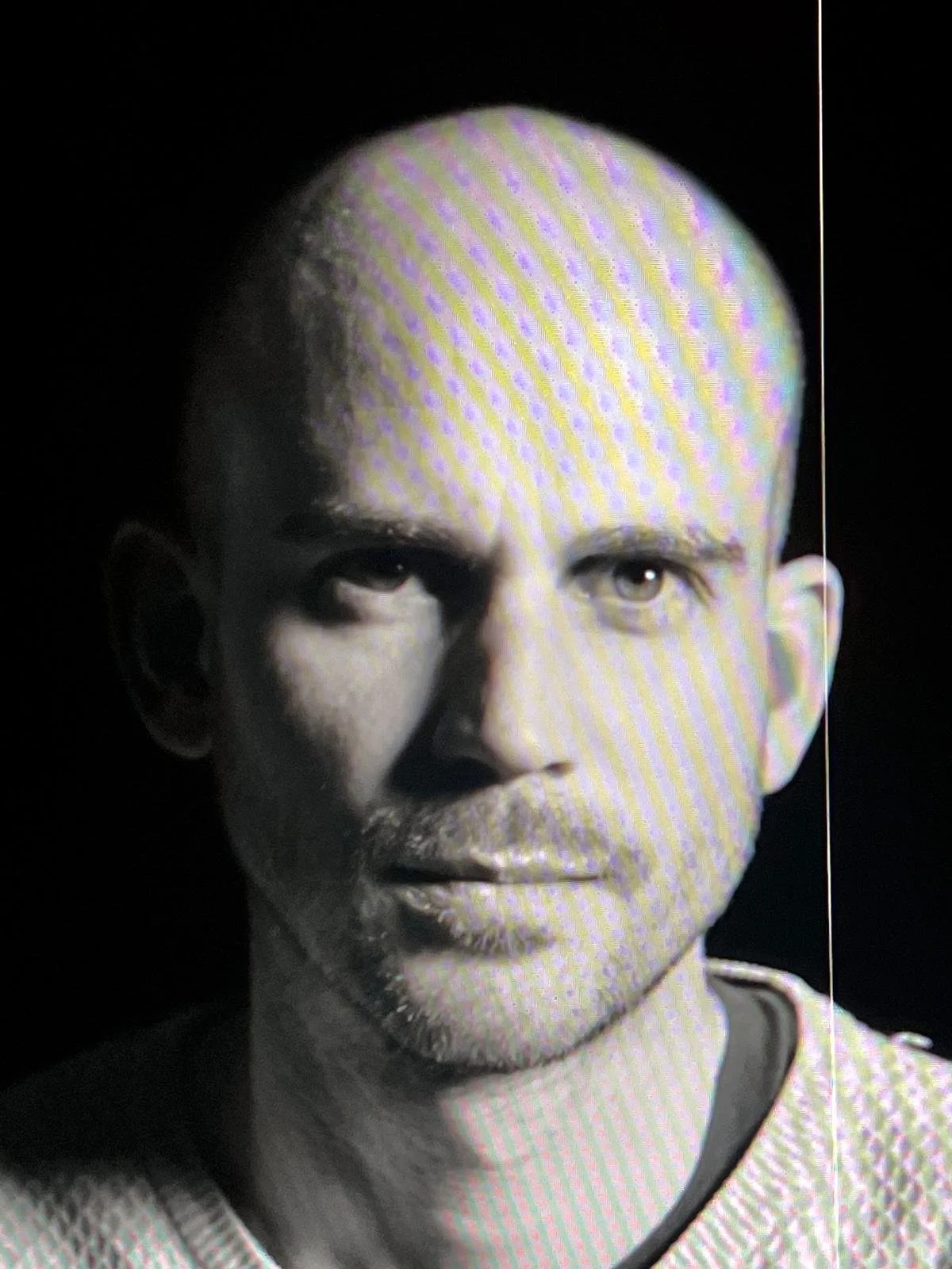 Une photo en noir et blanc d'un homme ayant un visage sérieux, avec une tête chauve, un regard perçant, et une barbe légère.