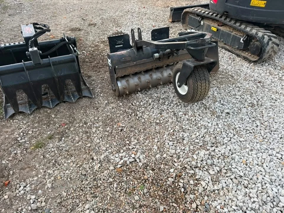 Heavy equipment attachments, including a trencher and toothed bucket, beside a tracked excavator on a gravel lot
