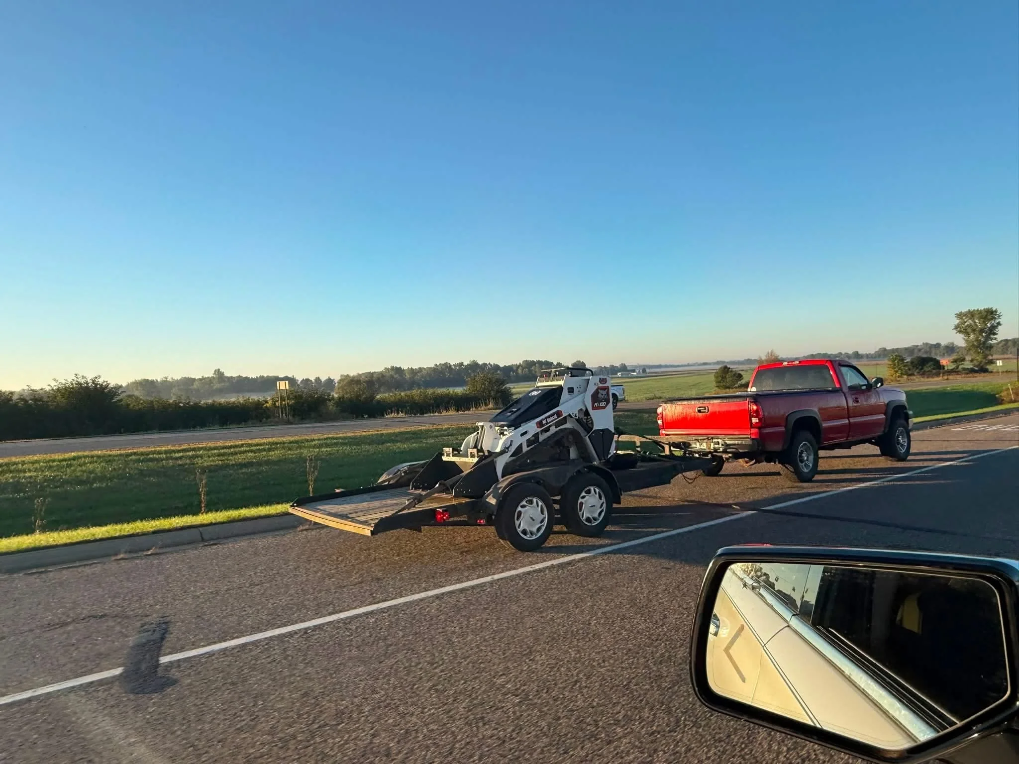 Red pickup truck towing a trailer with a compact white skid-steer loader on a rural highway.