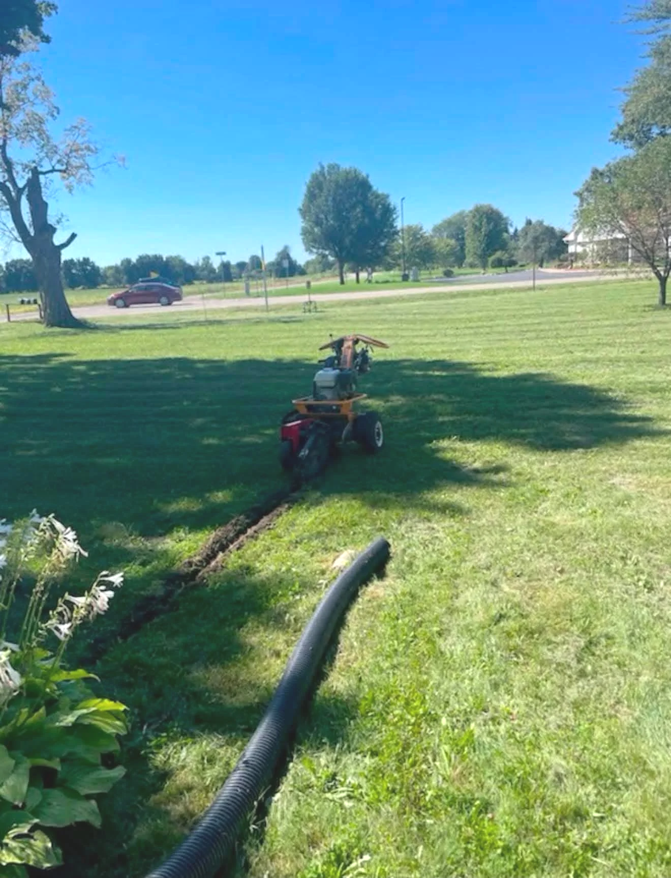 Lawn trenching machine digging a narrow trench across a grassy yard beside a large black corrugated pipe.