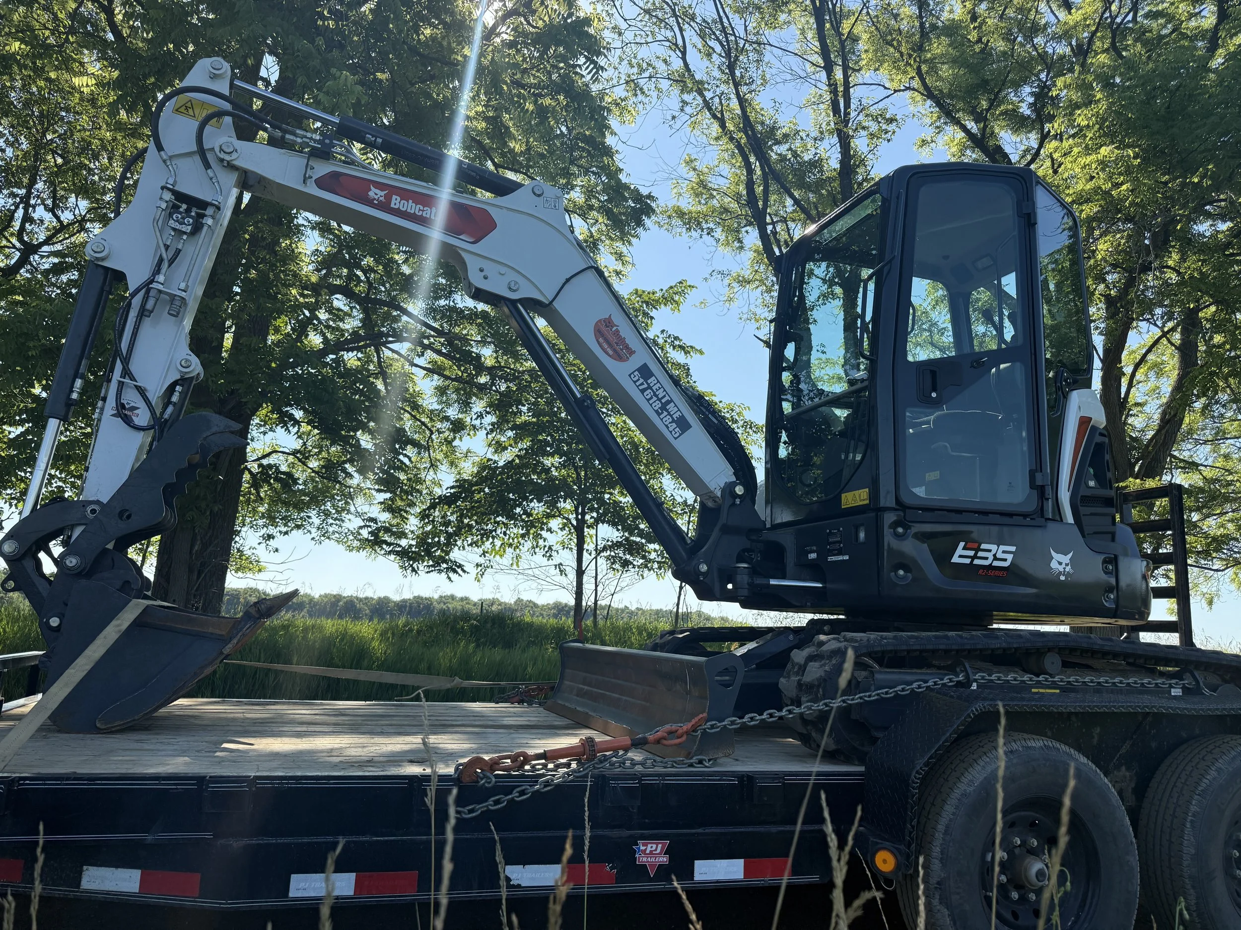A mini excavator on a flatbed trailer in an outdoor setting with trees and a clear sky.