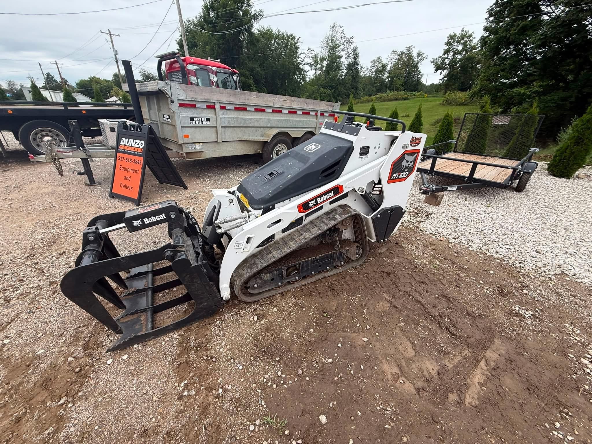 Bobcat MT100 compact track loader with a front grapple attachment beside a utility trailer and dump truck at an equipment rental yard