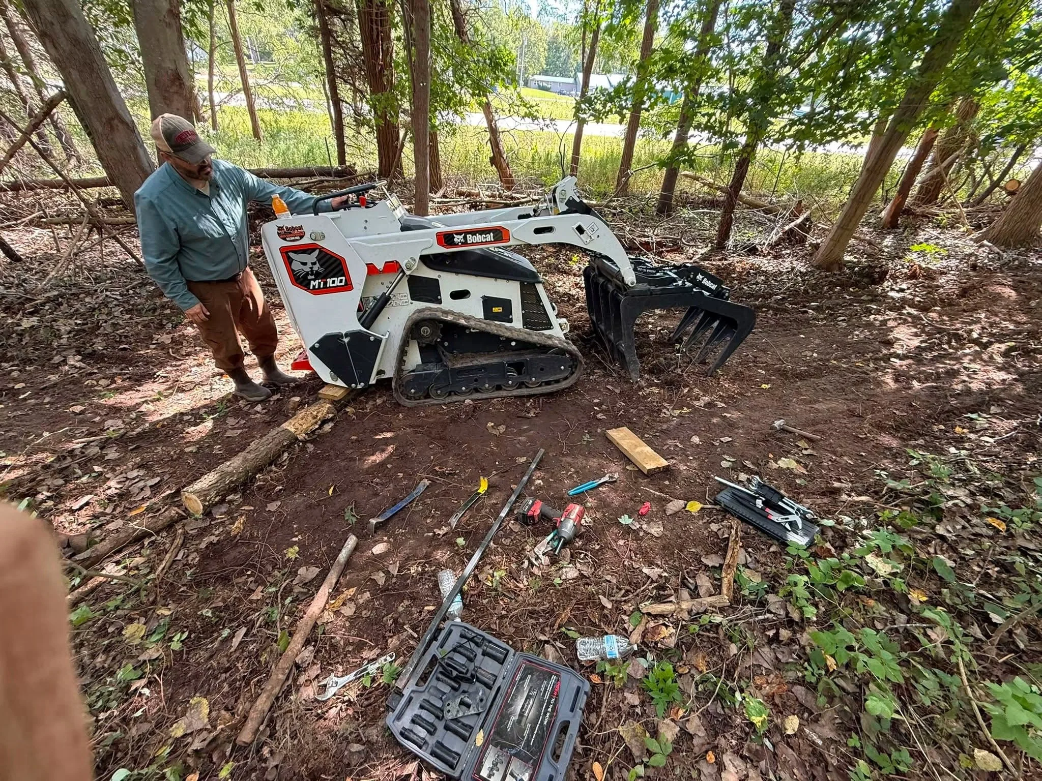 Man standing beside a Bobcat MT100 compact track loader with a grapple attachment in a wooded area, surrounded by tools and debris.