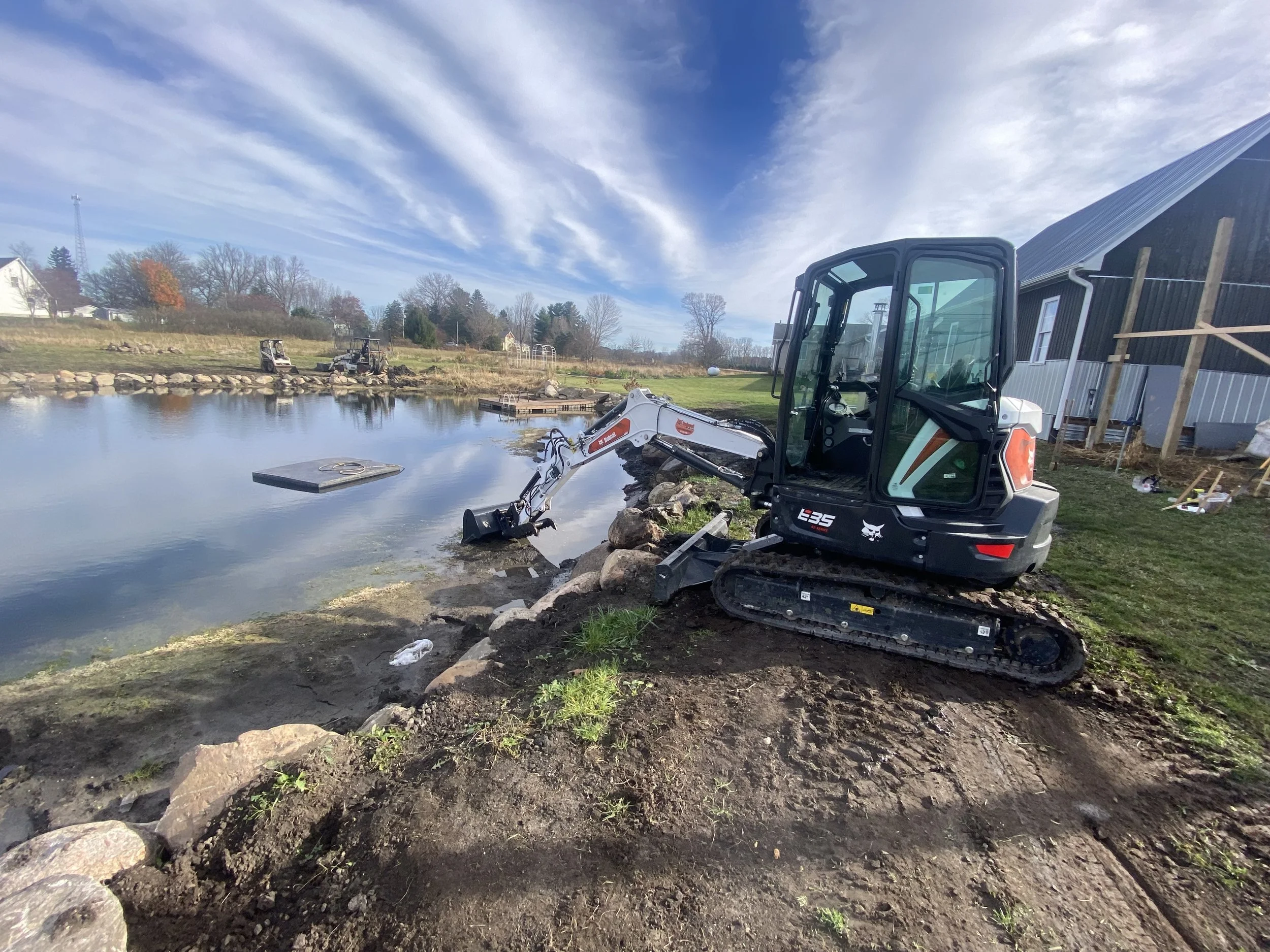 A small excavator near a body of water at a construction site on a sunny day with partly cloudy sky.