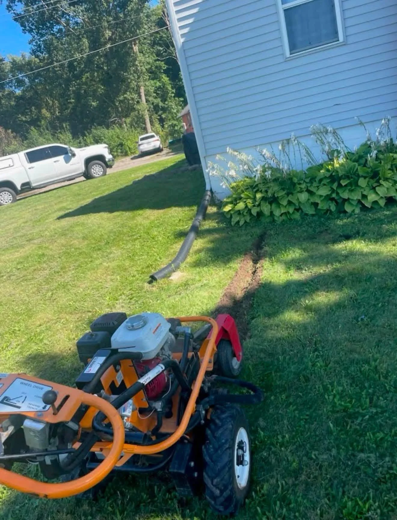 Orange walk-behind trenching machine beside a white-sided house, with a drainage hose extending across the lawn.