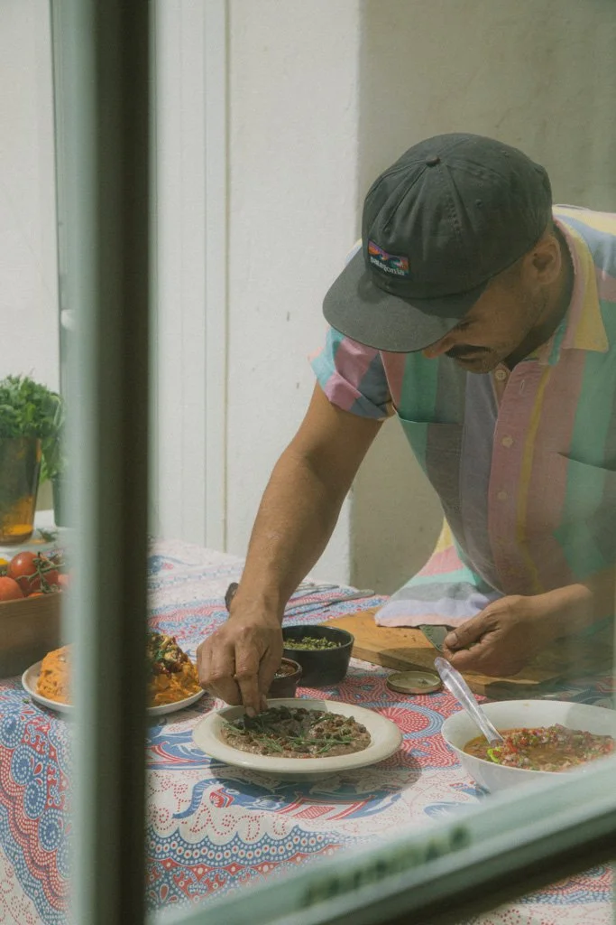 A man wearing a multicolored, striped shirt and a black Patagonia cap is reaching across a dining table with various dishes, including a pizza, a bowl of salad, and a plate of pasta, seen through a window.