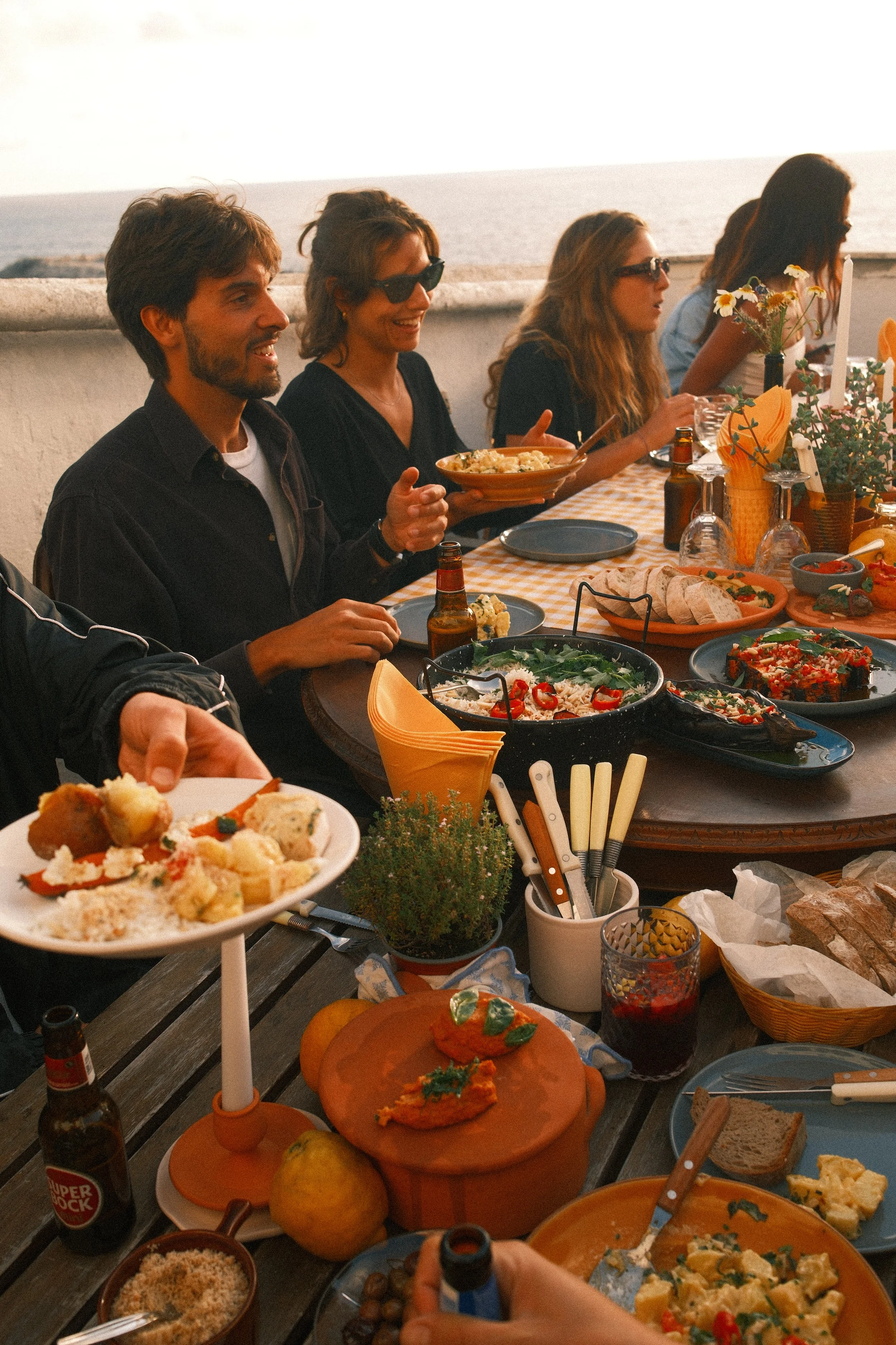 People enjoying a meal at a table outdoors by the ocean, with various dishes including salads, pizza, bread, and drinks, as the sunset or sunrise casts a warm glow.