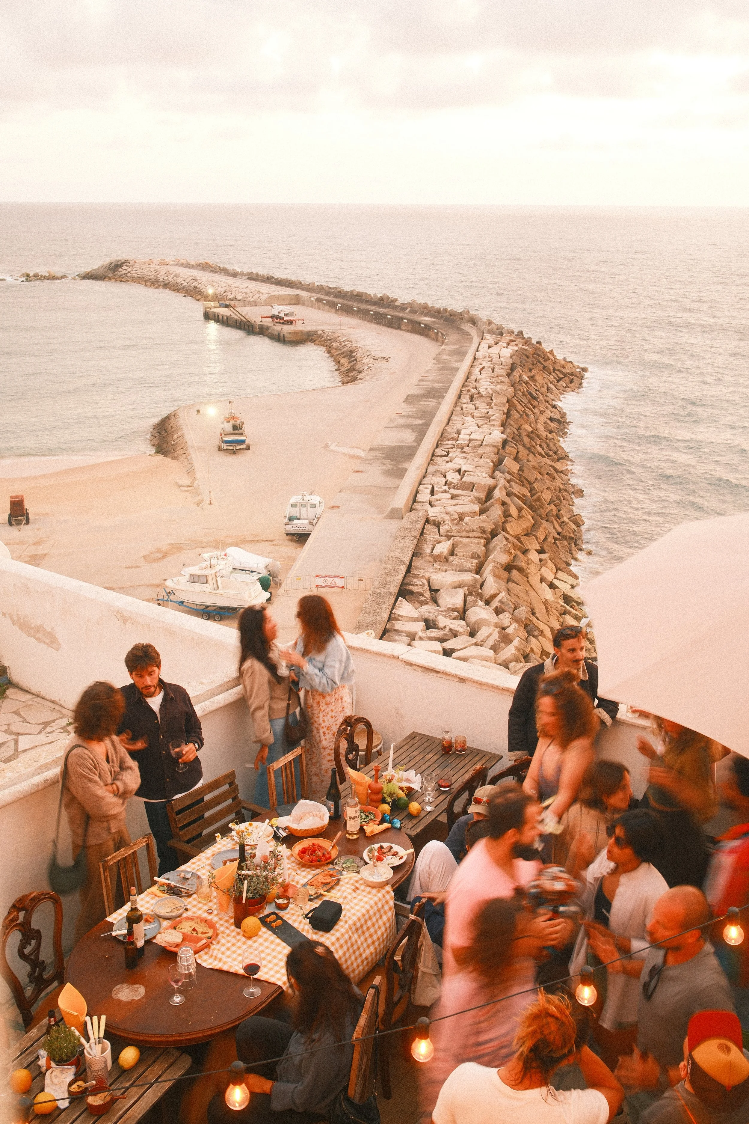 Group of people socializing and dining on a terrace with a view of the ocean, with a pier extending into the water and a table filled with food.