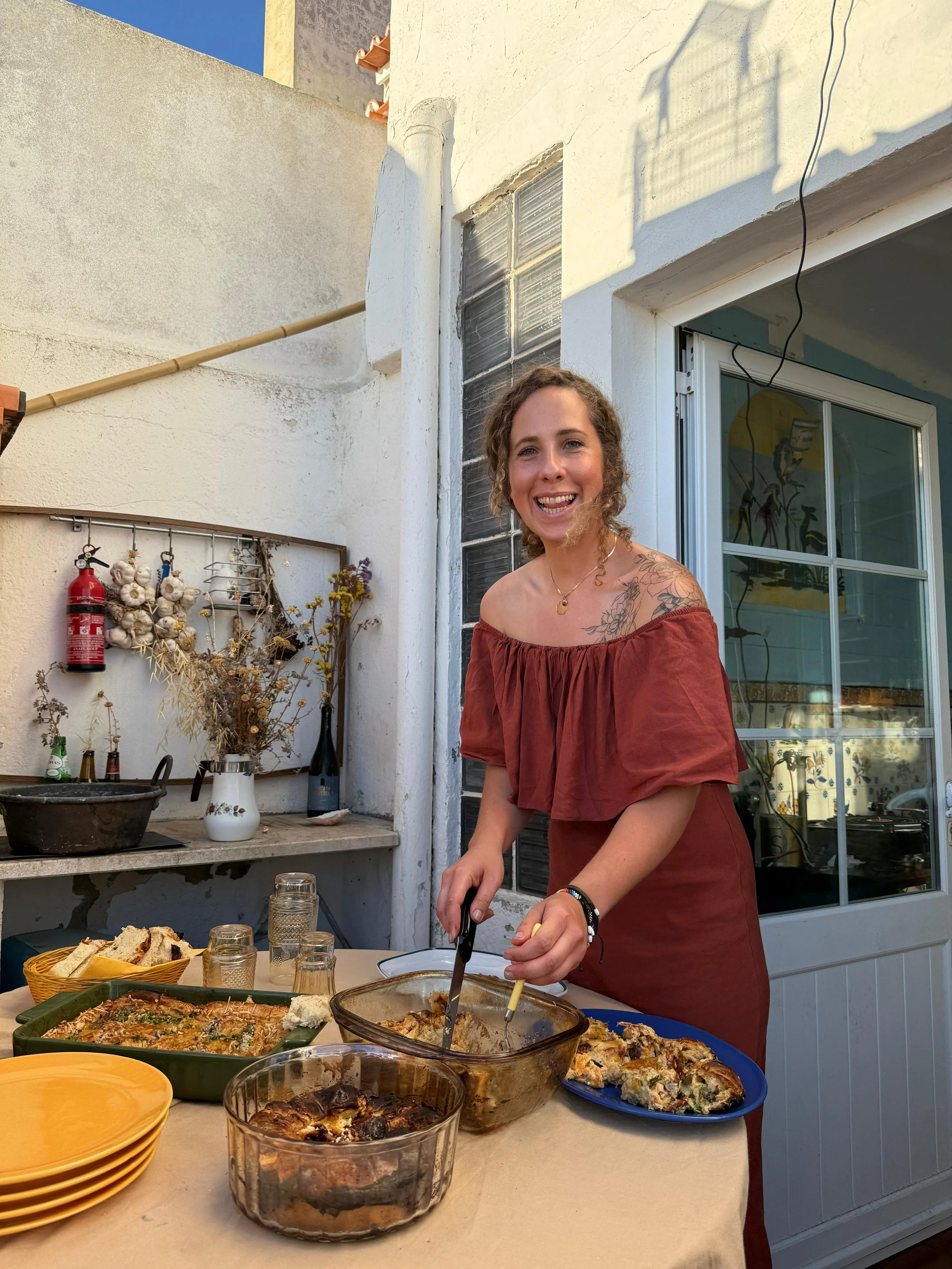 A woman with curly hair and tattoos on her shoulders is serving herself food at an outdoor gathering. There are various dishes on the table, including baked casseroles and sliced bread. The setting appears to be a patio with sunlight coming in, and there are decorations and kitchen items on a shelf behind her.