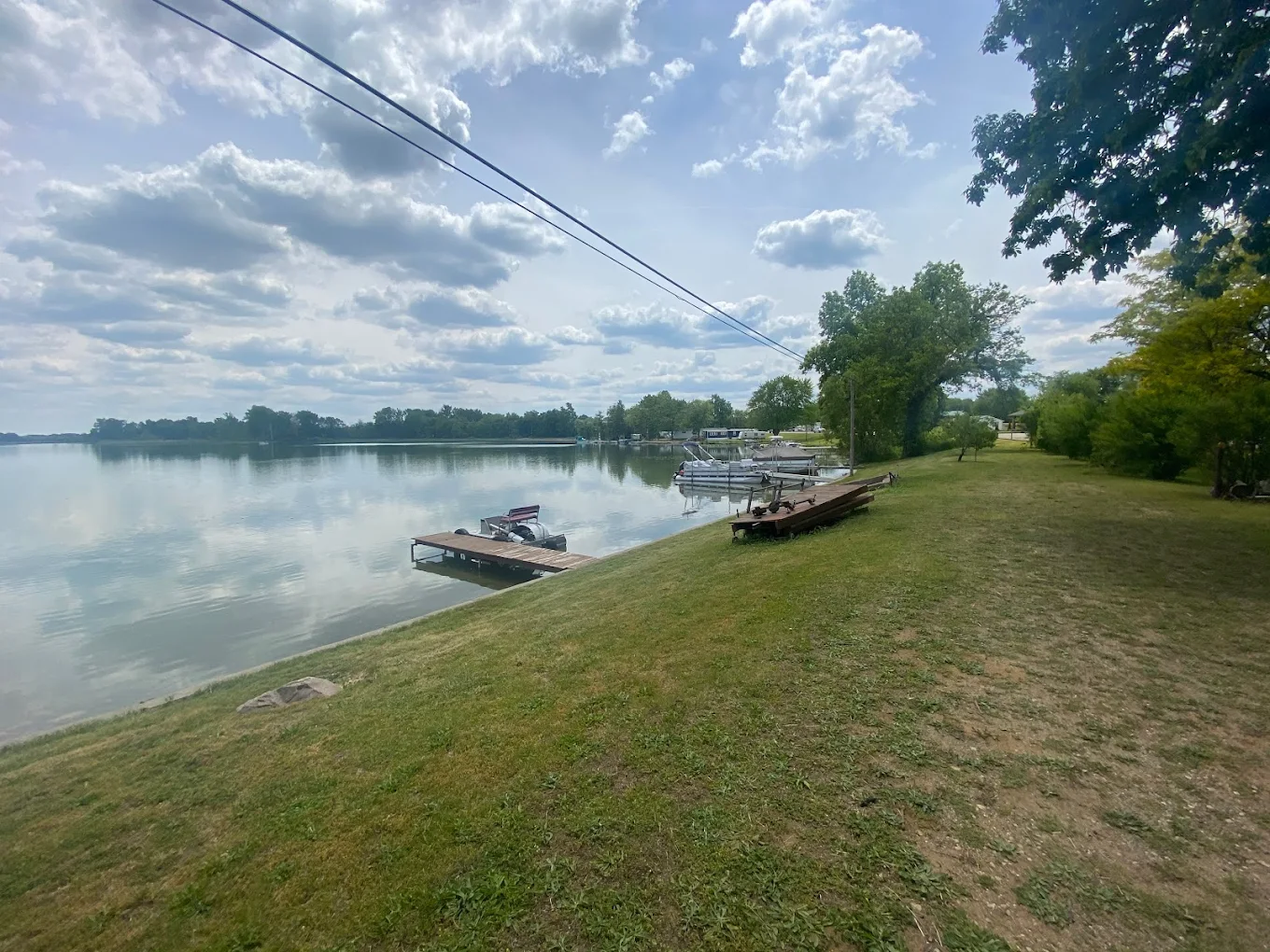 A peaceful lakeside scene with boats docked along the shoreline, grass, trees, and a partly cloudy sky reflected in calm water.