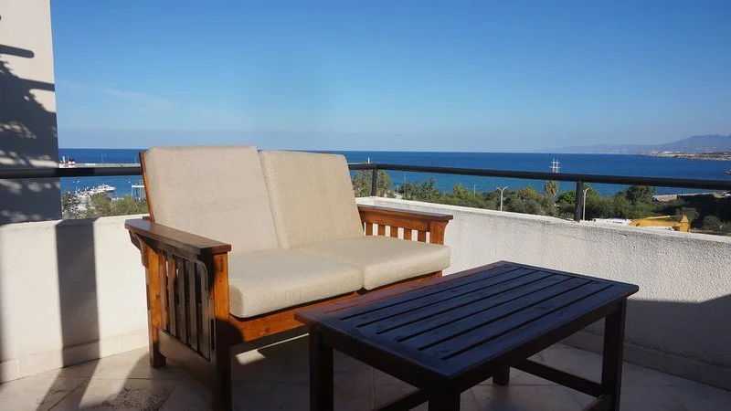 Outdoor balcony with a wooden and fabric sofa and a matching wooden table overlooking the ocean and distant shoreline under a clear blue sky.