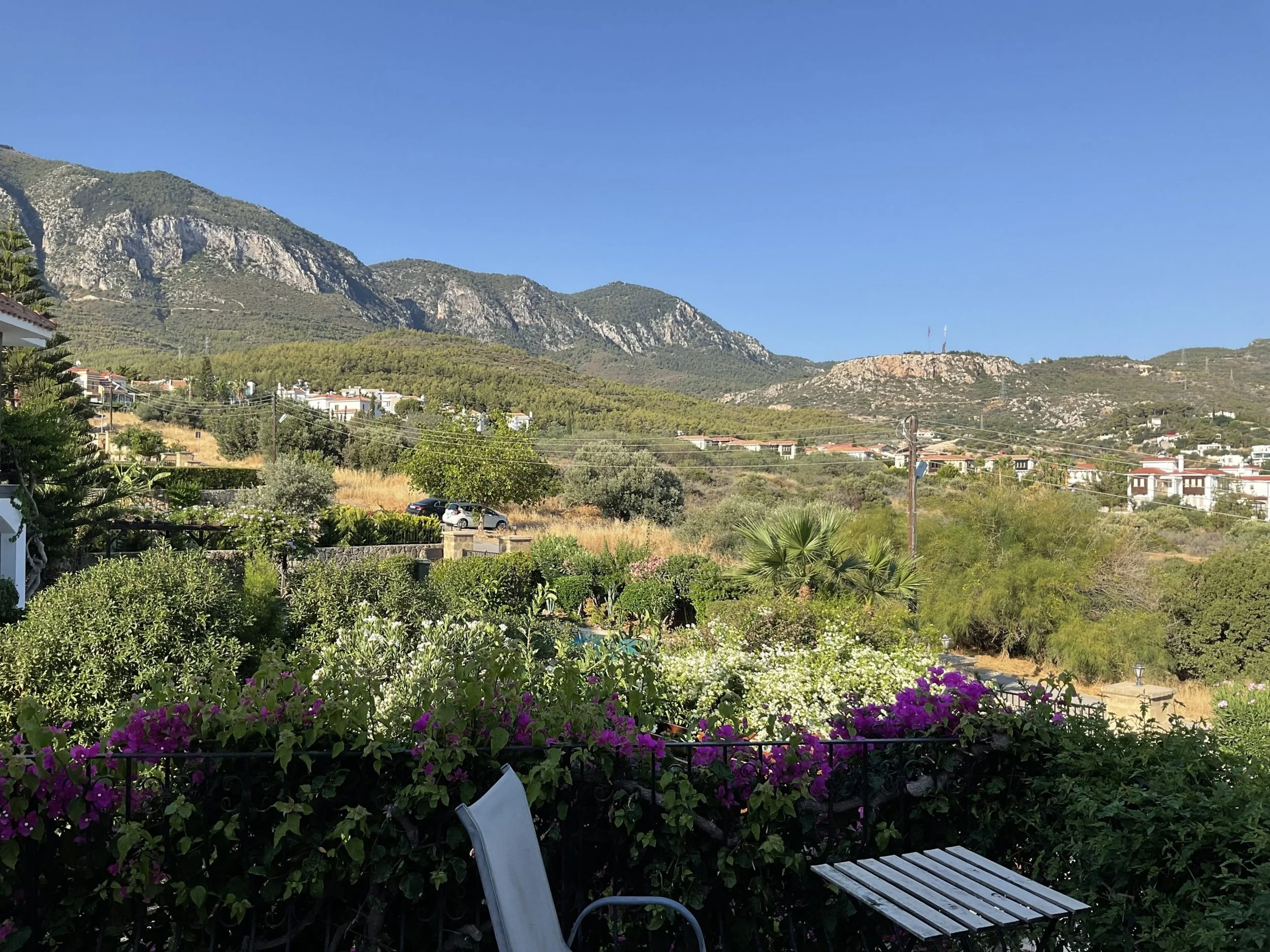 Scenic view of a garden with flowering bushes and trees, overlooking a hilly landscape with white houses and mountains in the background under a clear blue sky.