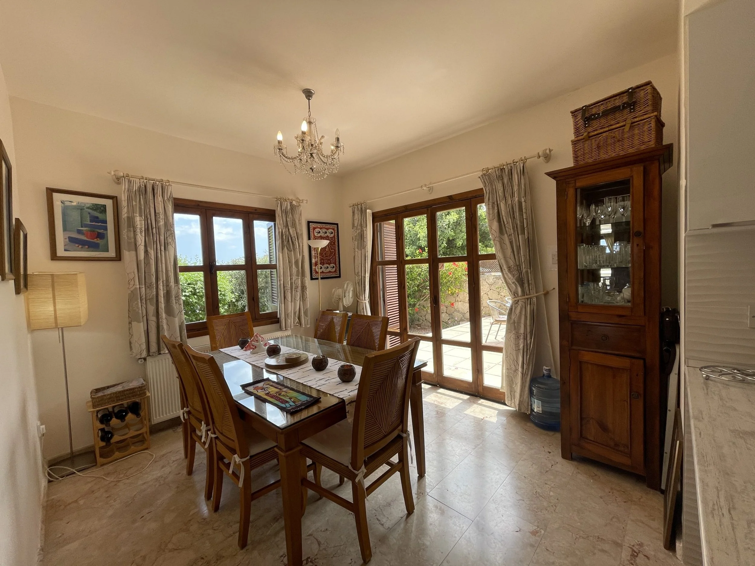 Dining room with wooden table and six chairs, chandelier, window and glass door leading outside, curtains, cabinet with glassware, water jug, and decorative items on the table.
